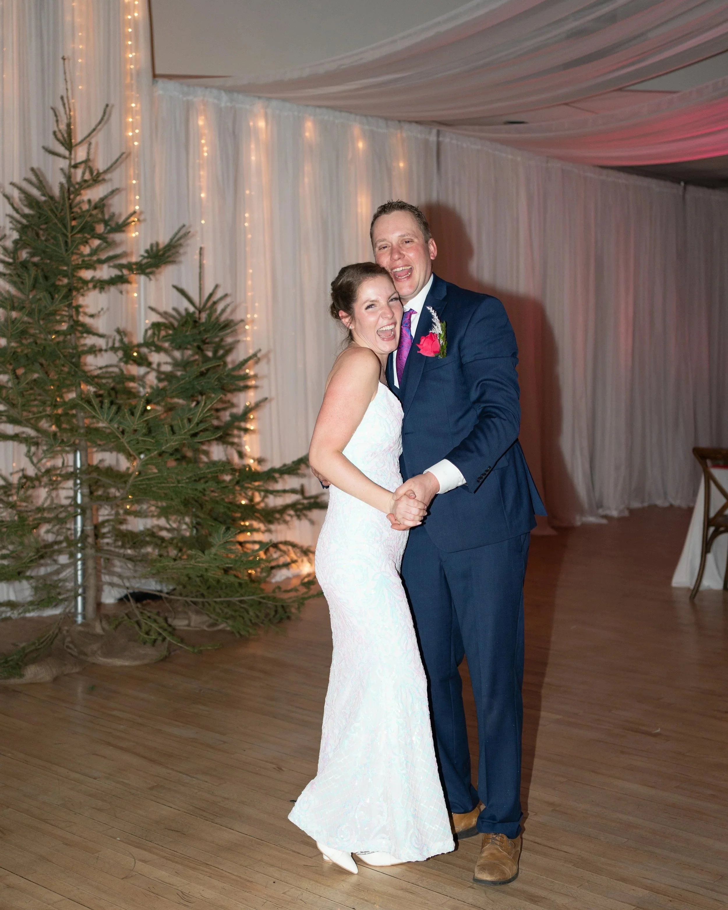 A happy newlywed couple dancing at their wedding reception, with a Christmas tree and string lights in the background. Wedding in Pembroke, Ontario