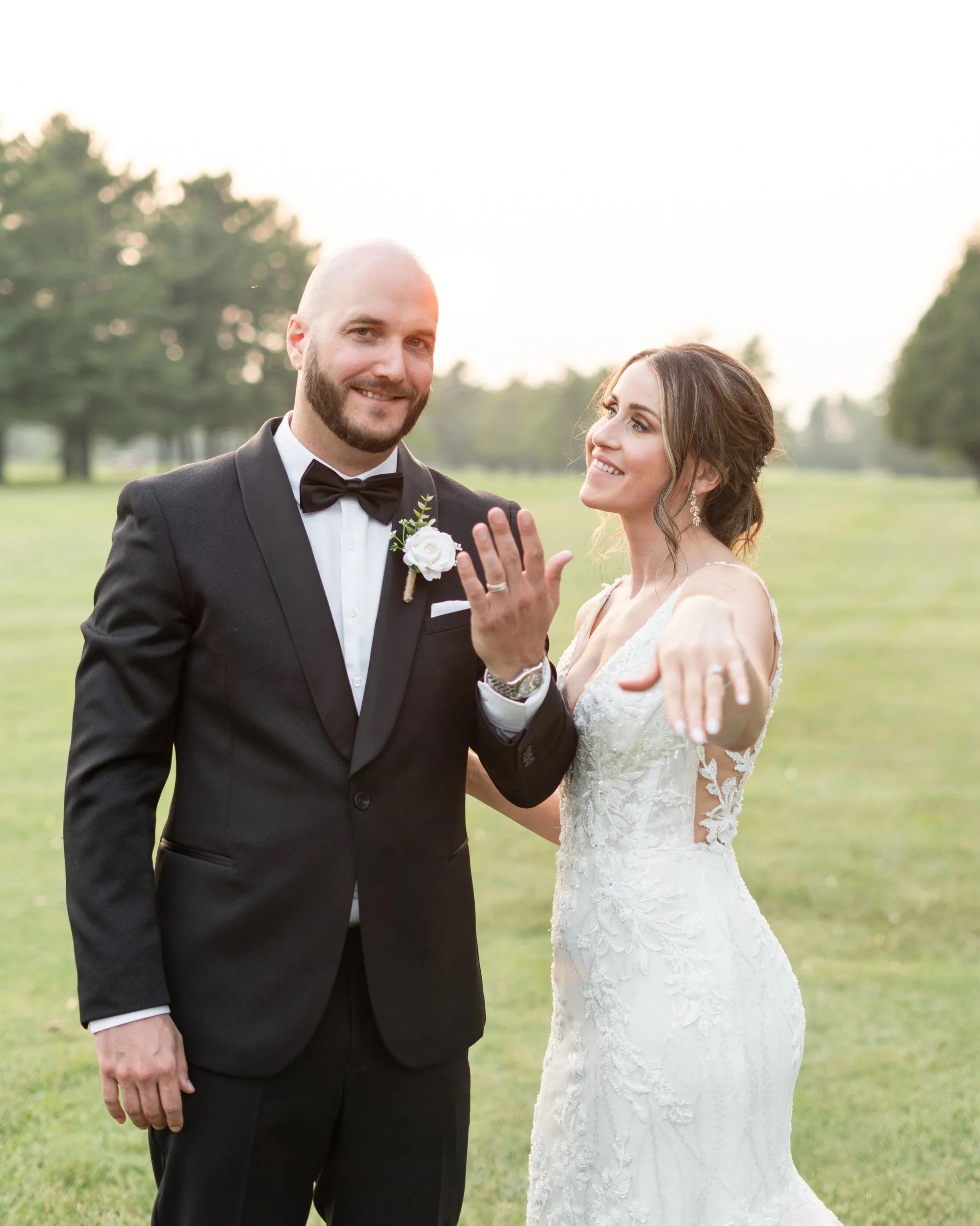 A bride and groom in wedding attire outdoors, with the groom displaying a wedding ring and the bride smiling and pointing. Pembroke Golf Club, Ontario