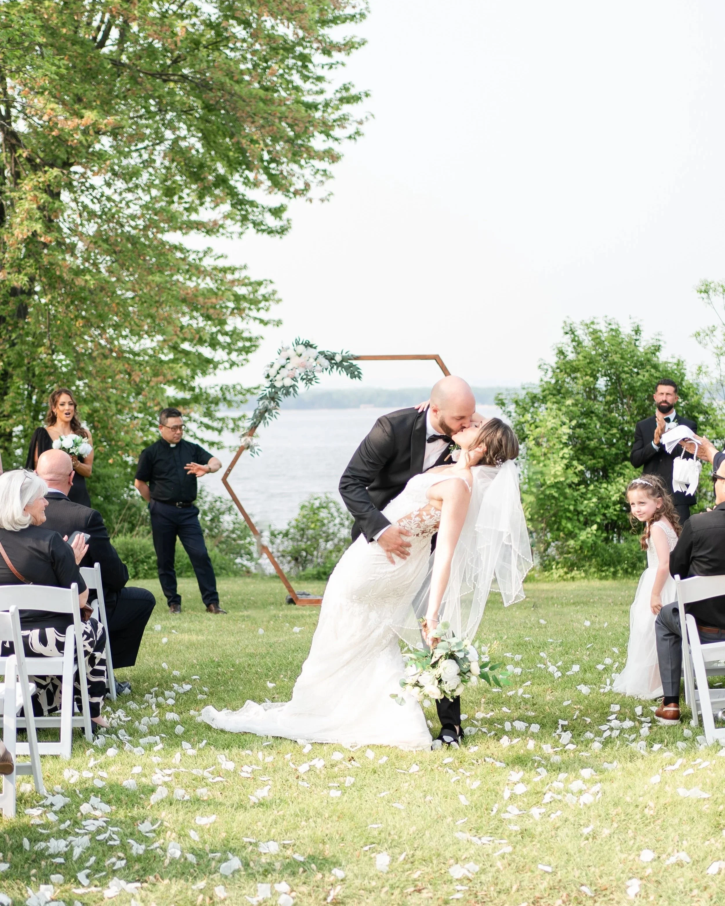 A wedding ceremony outdoors by a lake with a bride and groom sharing a kiss, surrounded by guests and floral decor. Pembroke Golf Club, Ontario