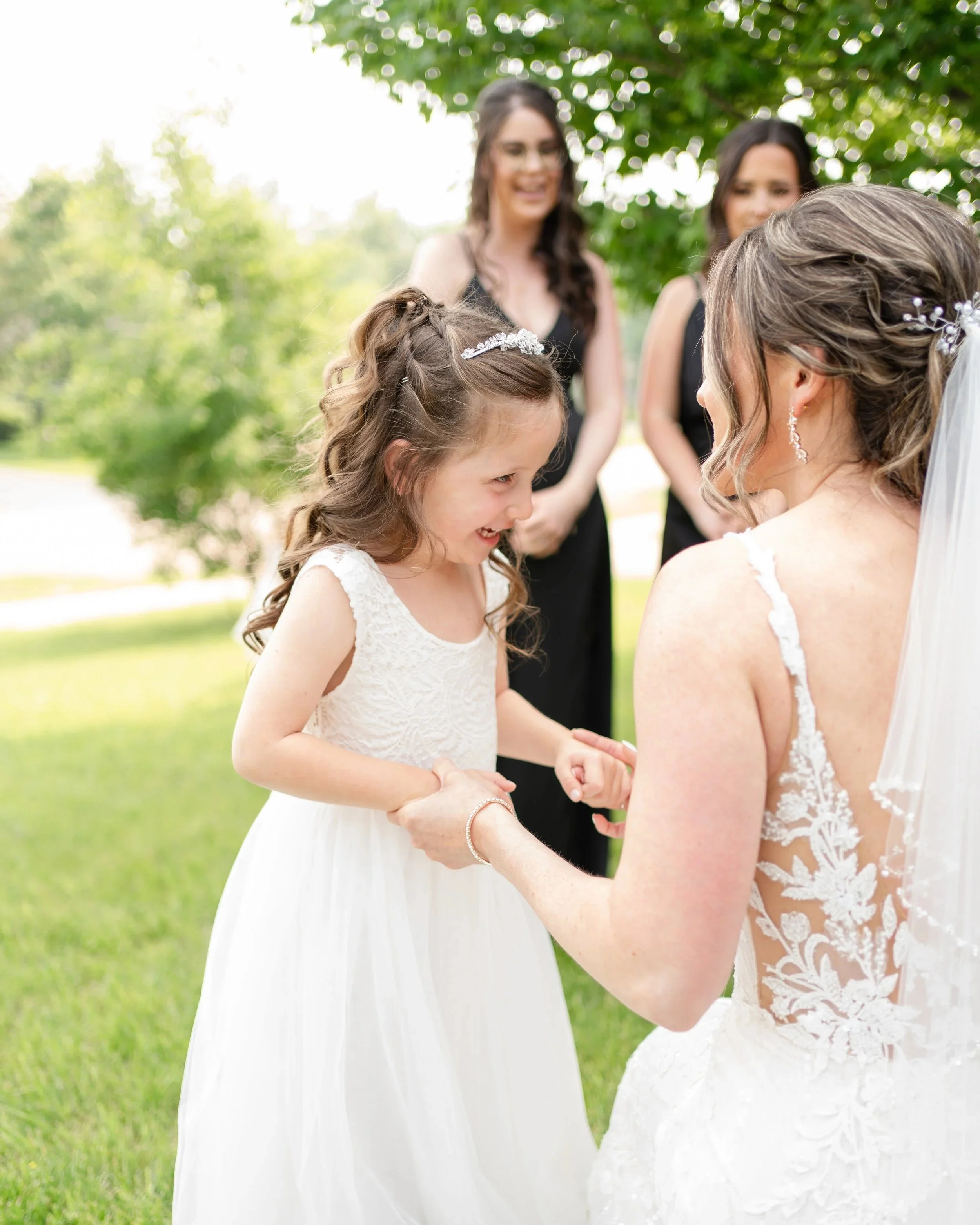 A young girl in a white dress smiling and holding hands with a woman in a wedding dress during an outdoor wedding ceremony, with three women in black dresses in the background.