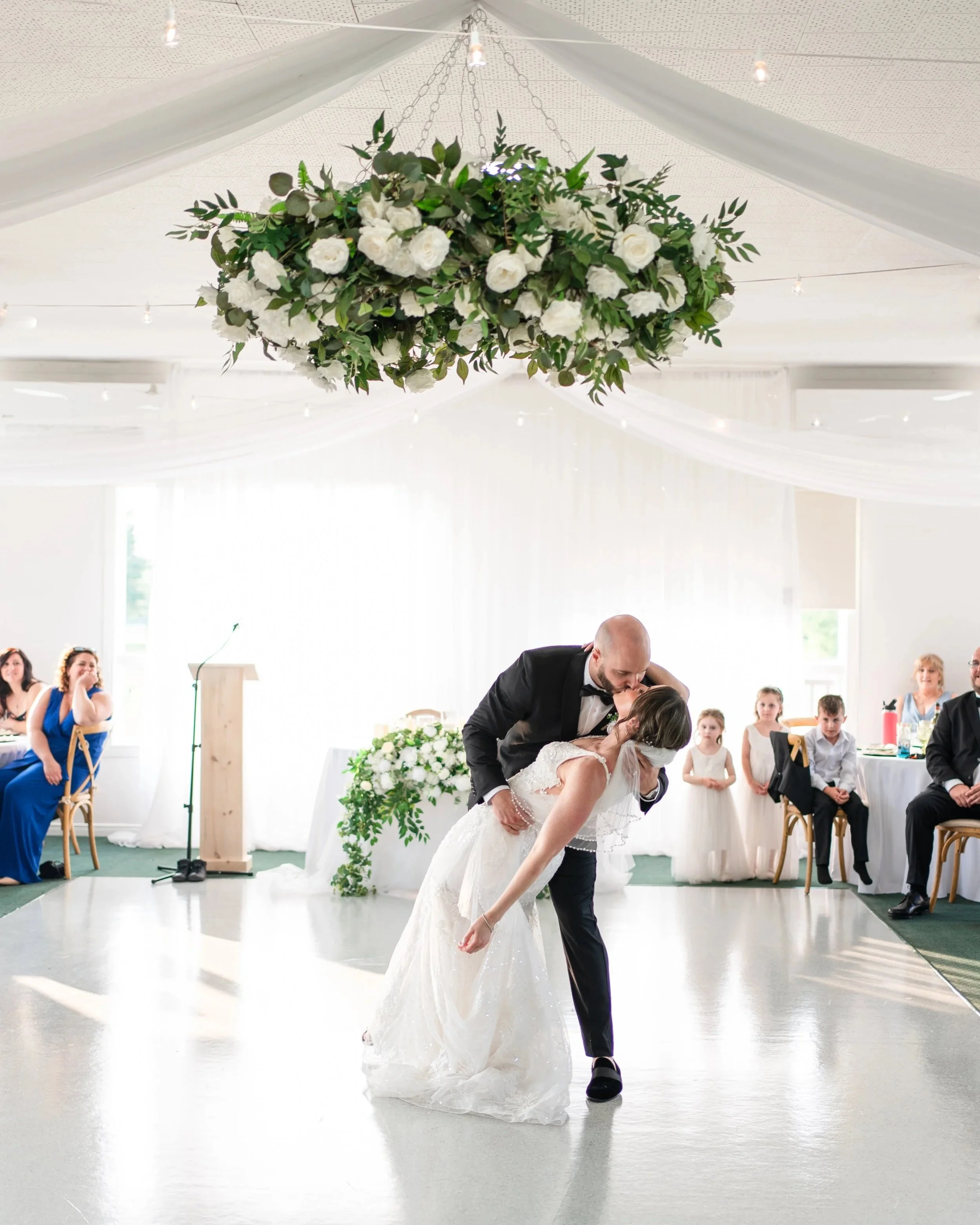 A newlywed couple sharing a kiss during their wedding dance in a decorated reception hall with seated guests watching. Pembroke Golf Club, Ontario.