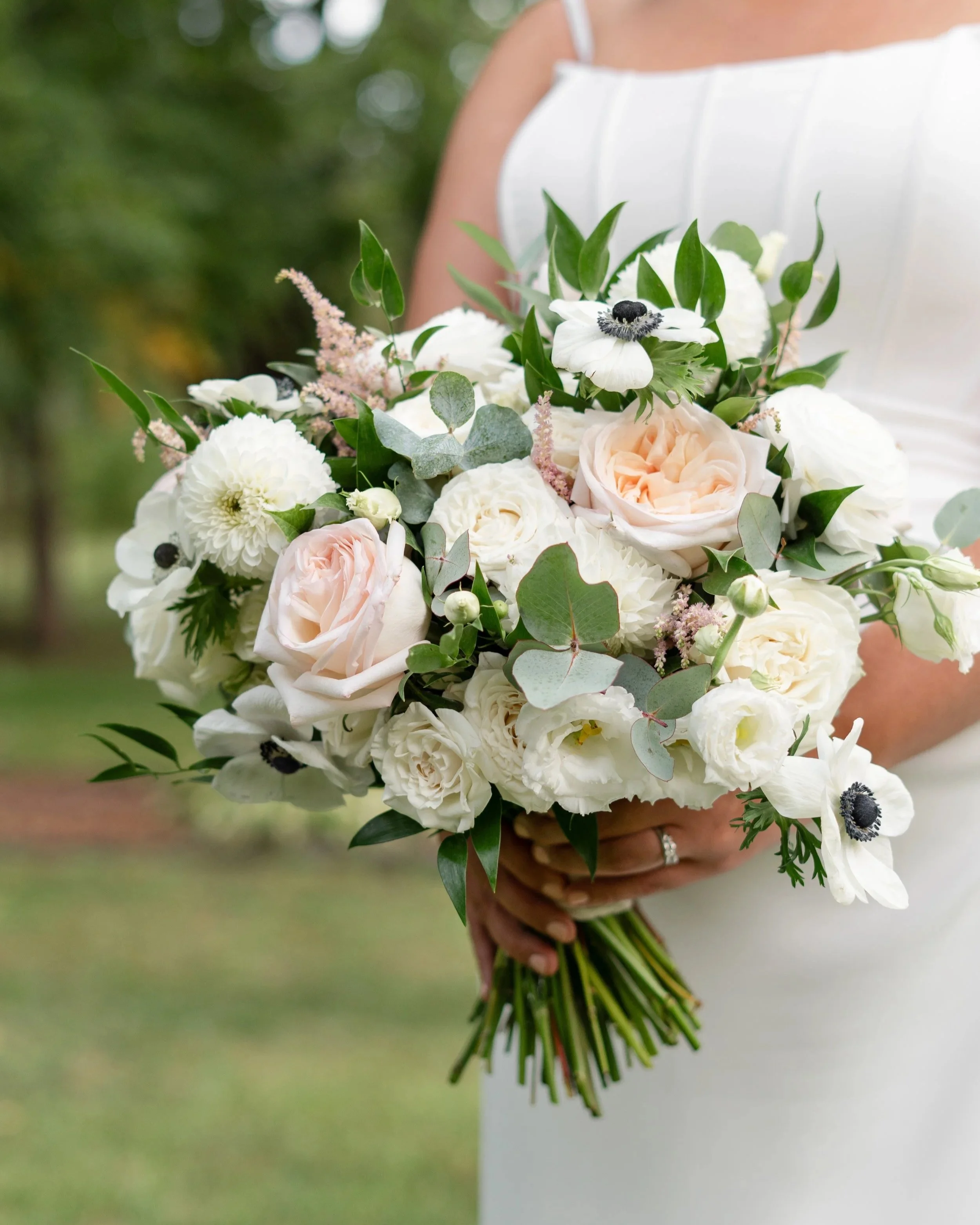 A person in a white dress holding a bouquet of white, blush pink, and green flowers in an outdoor setting. Pembroke Wedding.