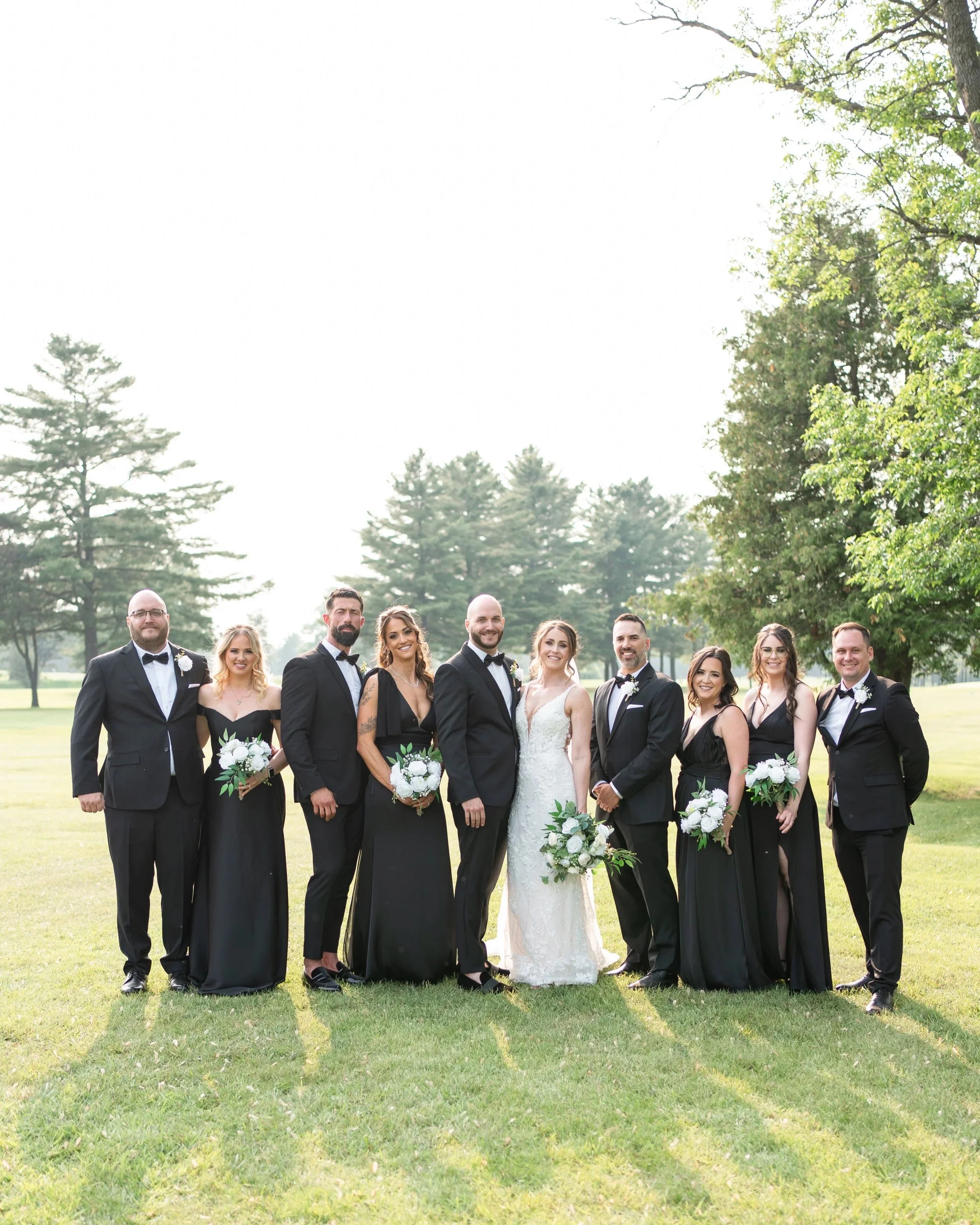 A wedding party with the bride and groom. The couple is surrounded by bridesmaids in black dresses holding bouquets, and groomsmen in black tuxedos in a park setting. Pemrboke golf club, ontario