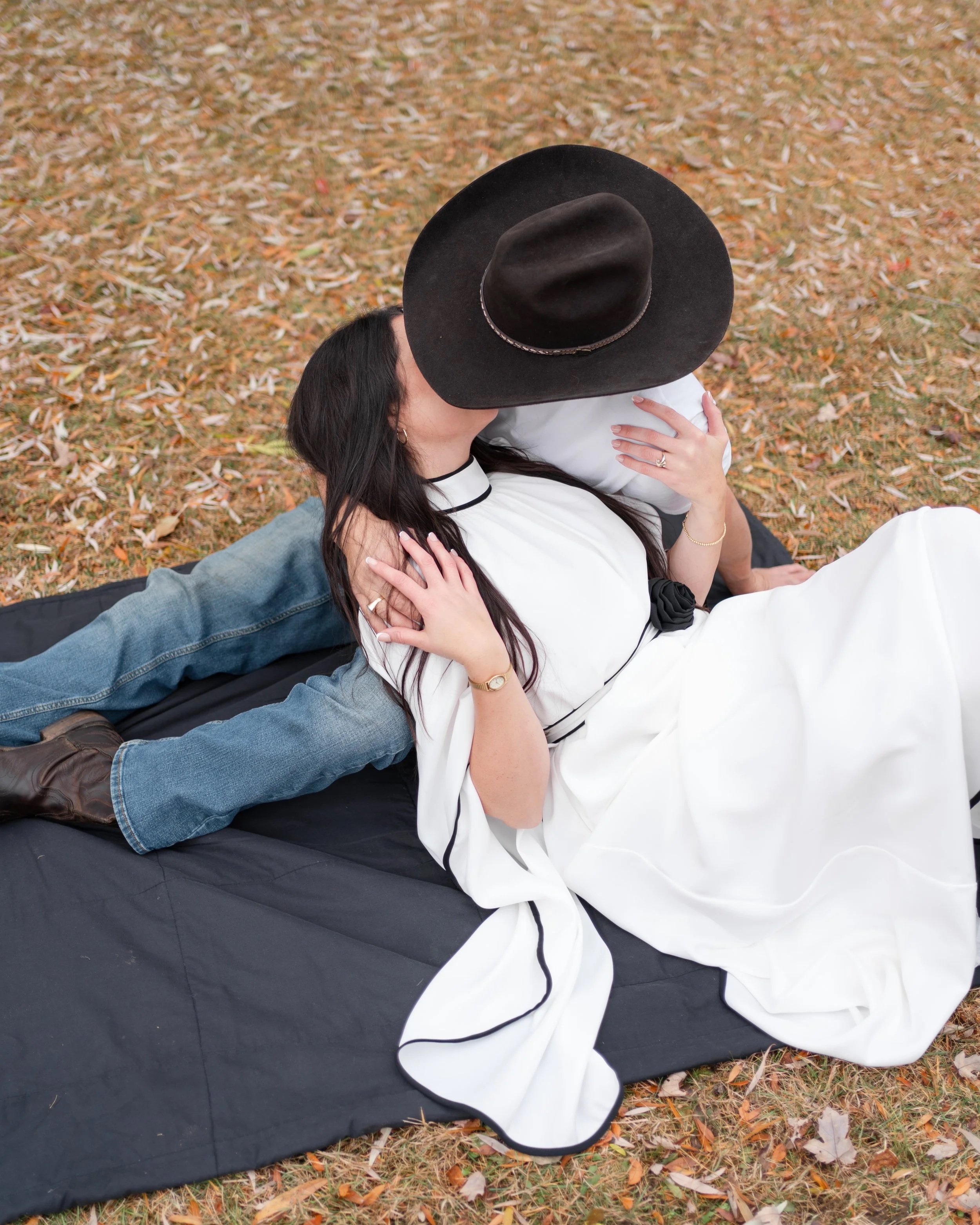 Fall engagement session in the Ottawa Valley. A woman in a white dress and large black hat is sitting on a black blanket on fallen leaves, embracing a person laying behind her, who is wearing jeans and brown boots.