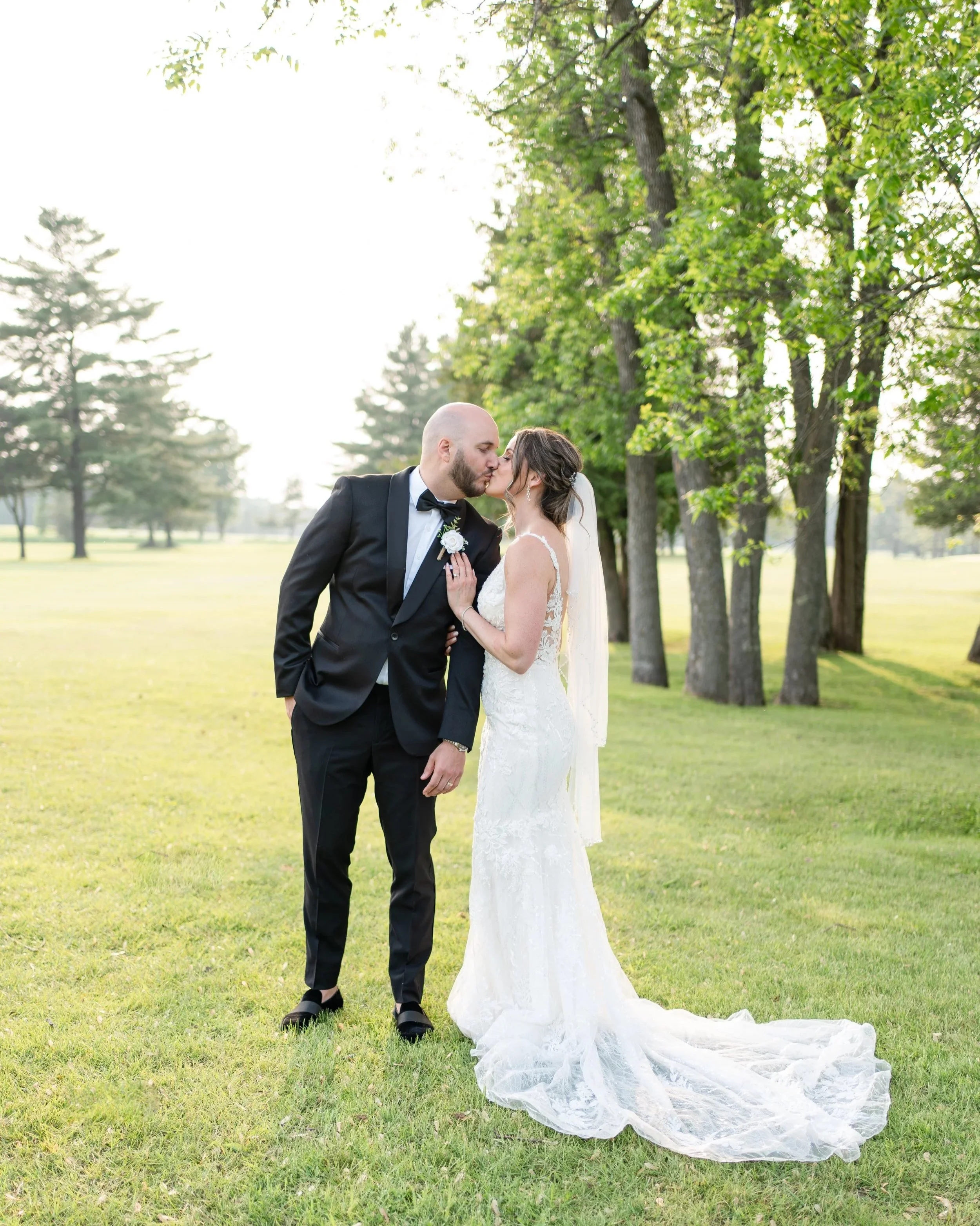 A bride and groom share a kiss outdoors on a sunny day in a park with green trees and grass. Wedding at Pembroke Golf Club, Ontario.