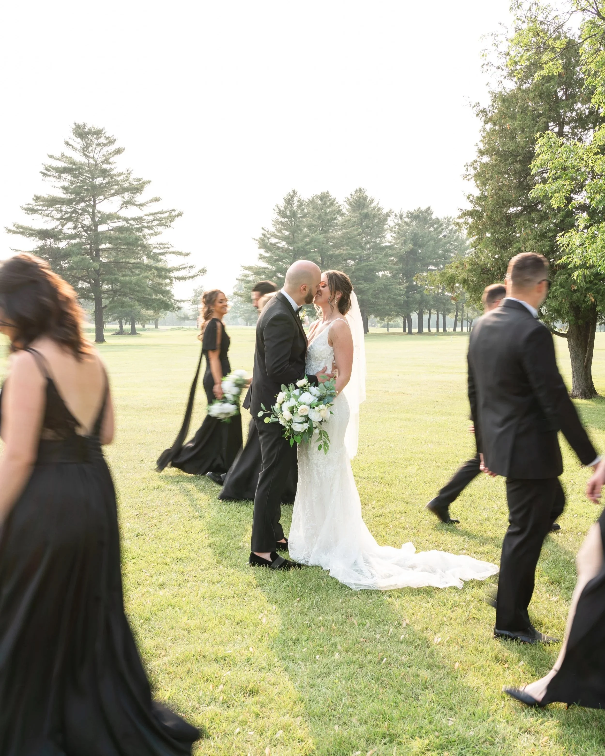 Bride and groom kissing at outdoor wedding on grassy field, surrounded by bridesmaids and groomsmen. Pembroke Golf Club, Ontario