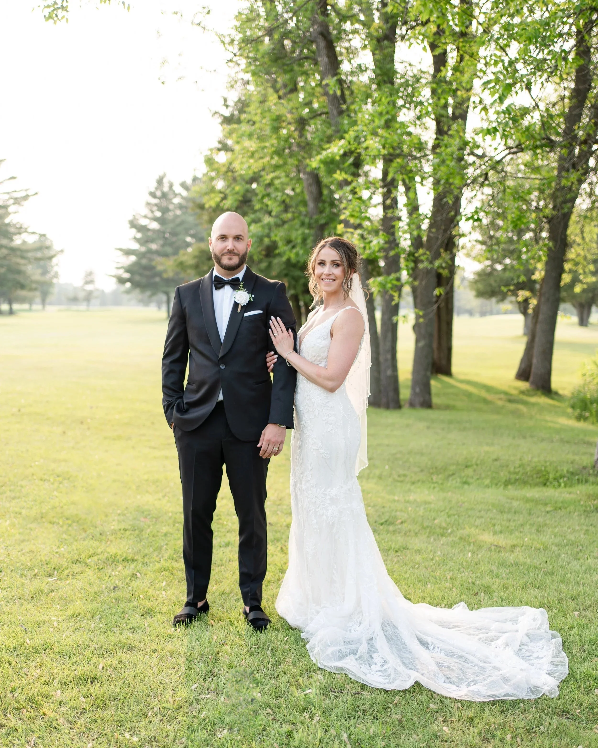 Married couple standing outdoors in front of trees with green leaves, smiling at the camera. The groom is wearing a black tuxedo with a bow tie and boutonniere, and the bride is in a white lace wedding gown with a veil. Pembroke Golf Club, ON