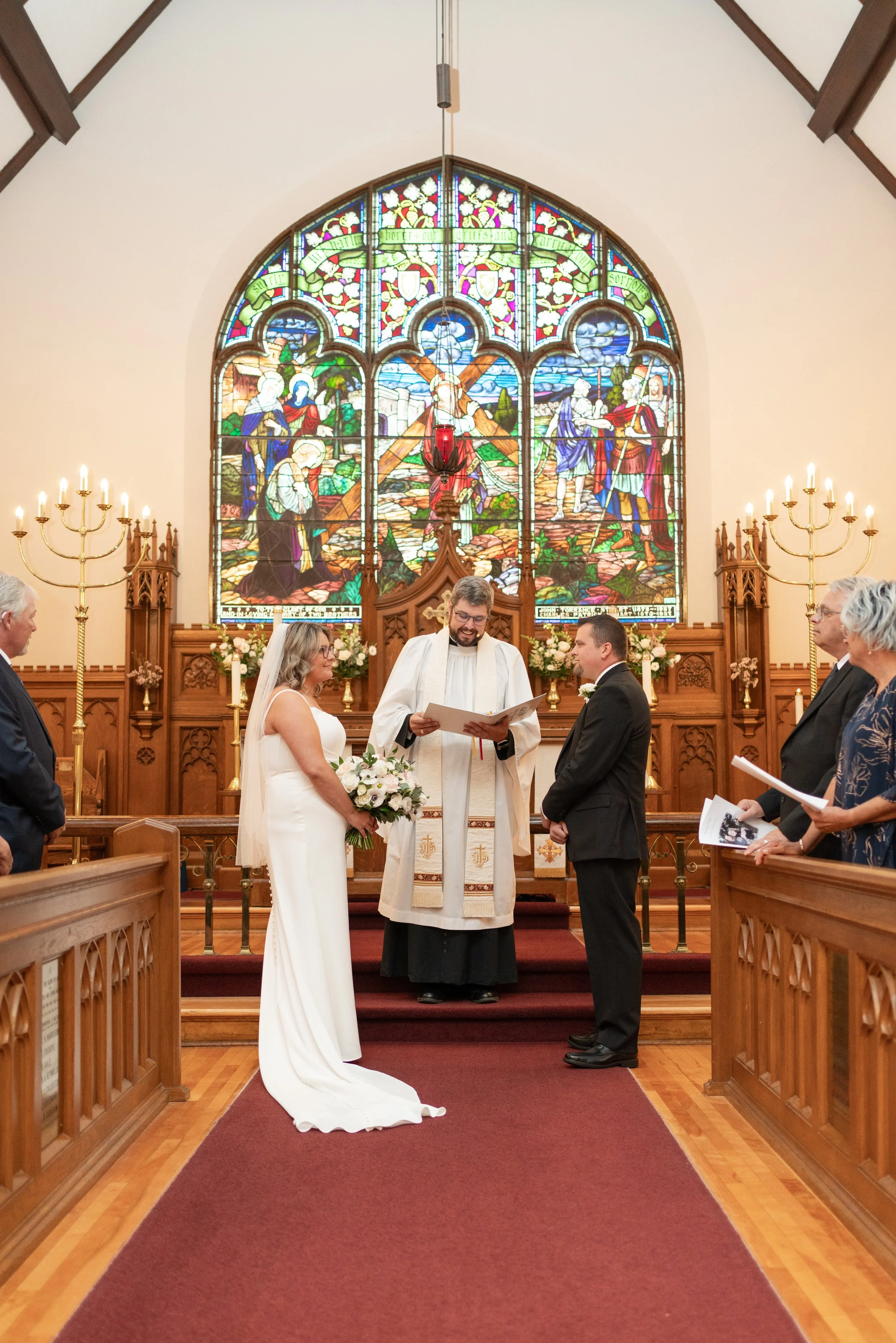 A wedding ceremony taking place inside a church with stained glass windows depicting religious scenes, where a bride and groom are standing before a priest, surrounded by guests. Church in Pembroke, Ontario