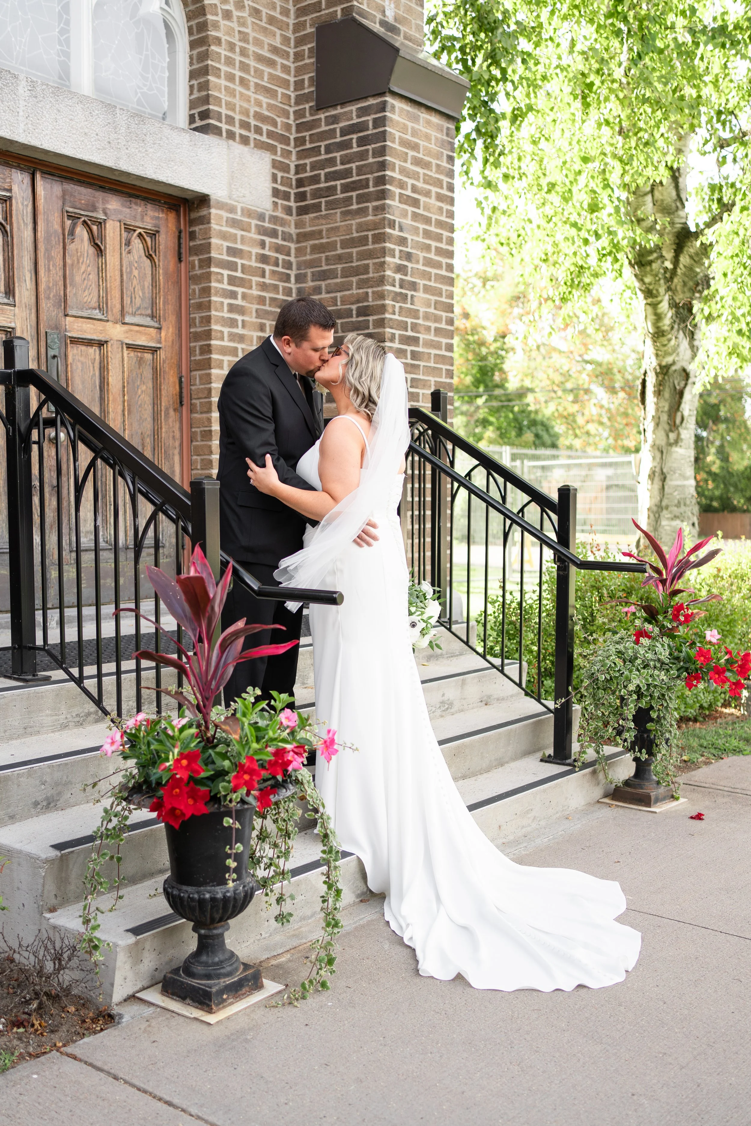 A bride and groom sharing a kiss on the steps of a church, surrounded by colorful flowers and greenery. Church in Pembroke, Ontario