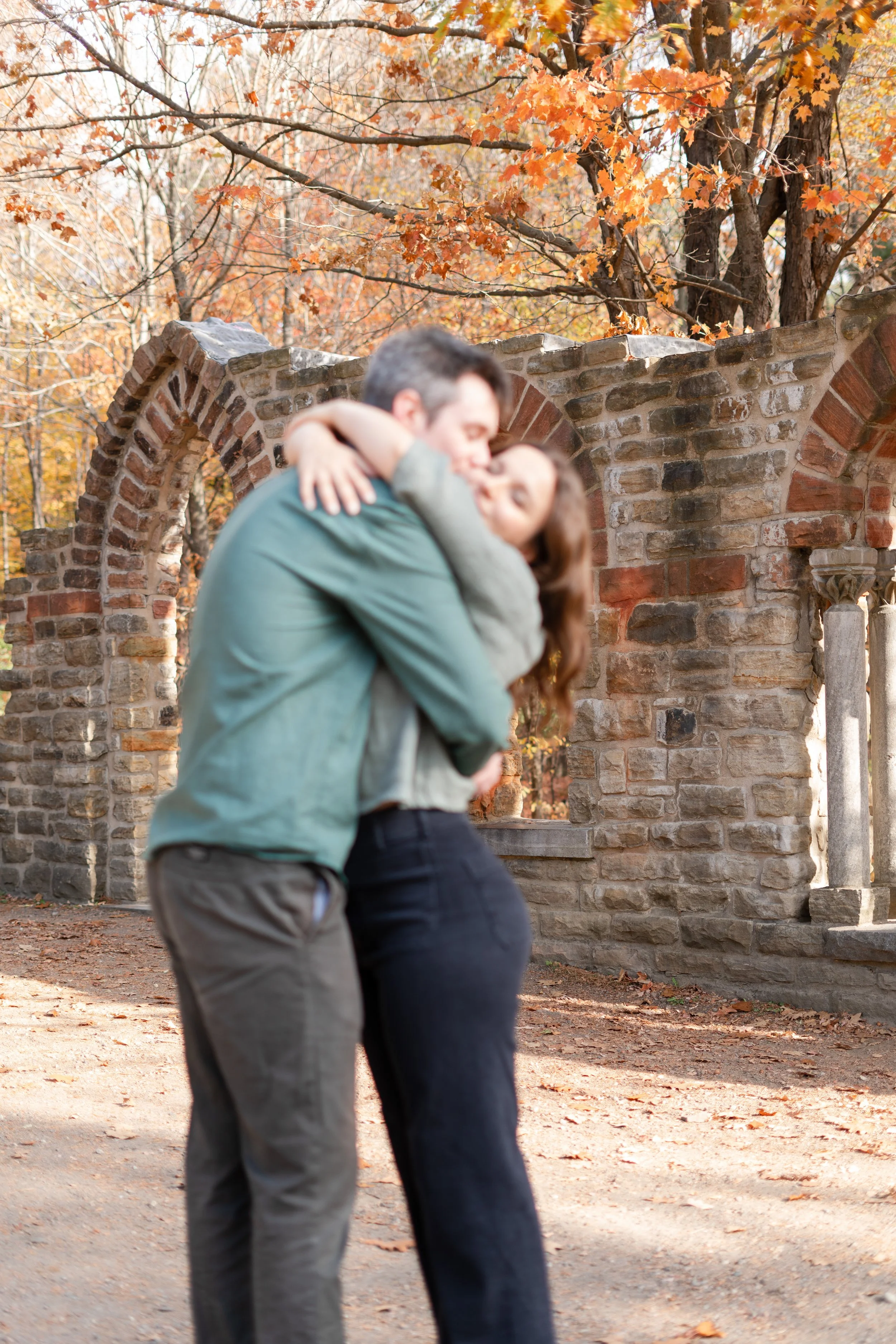 A couple hugging and kissing outdoors with autumn trees and a brick wall in the background.