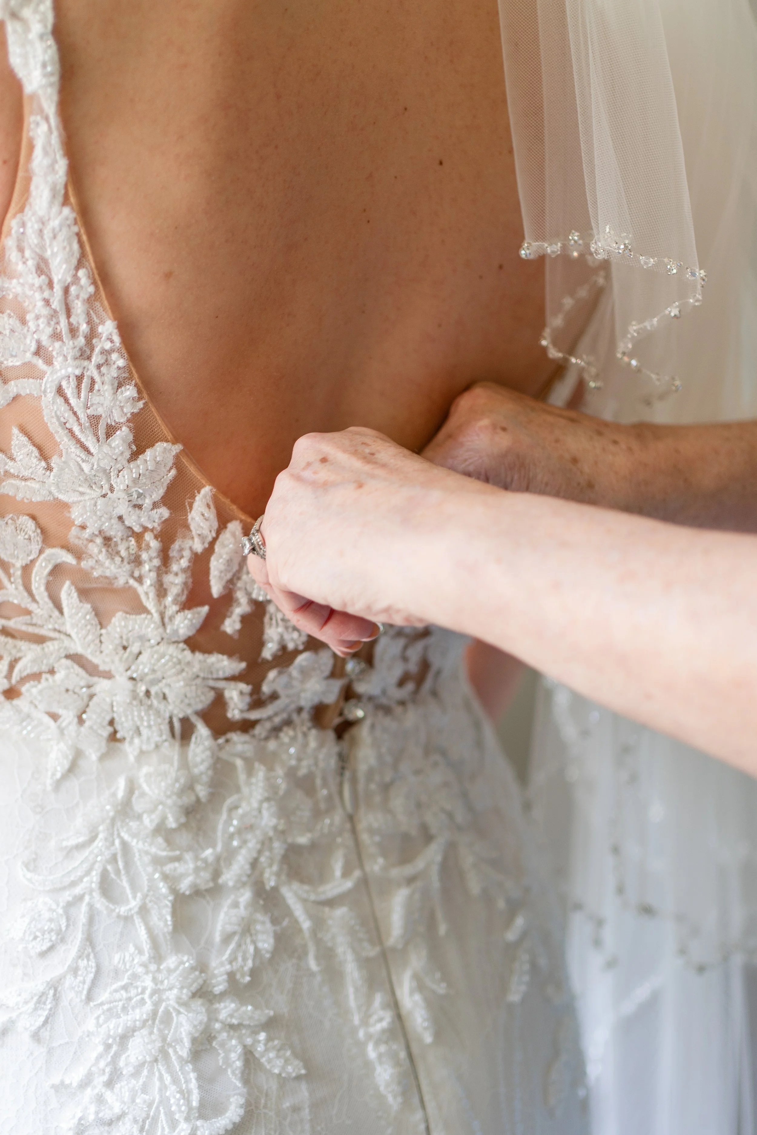 Close-up of a bride fastening her wedding dress, featuring intricate lace and embroidery details, with part of her veil visible.