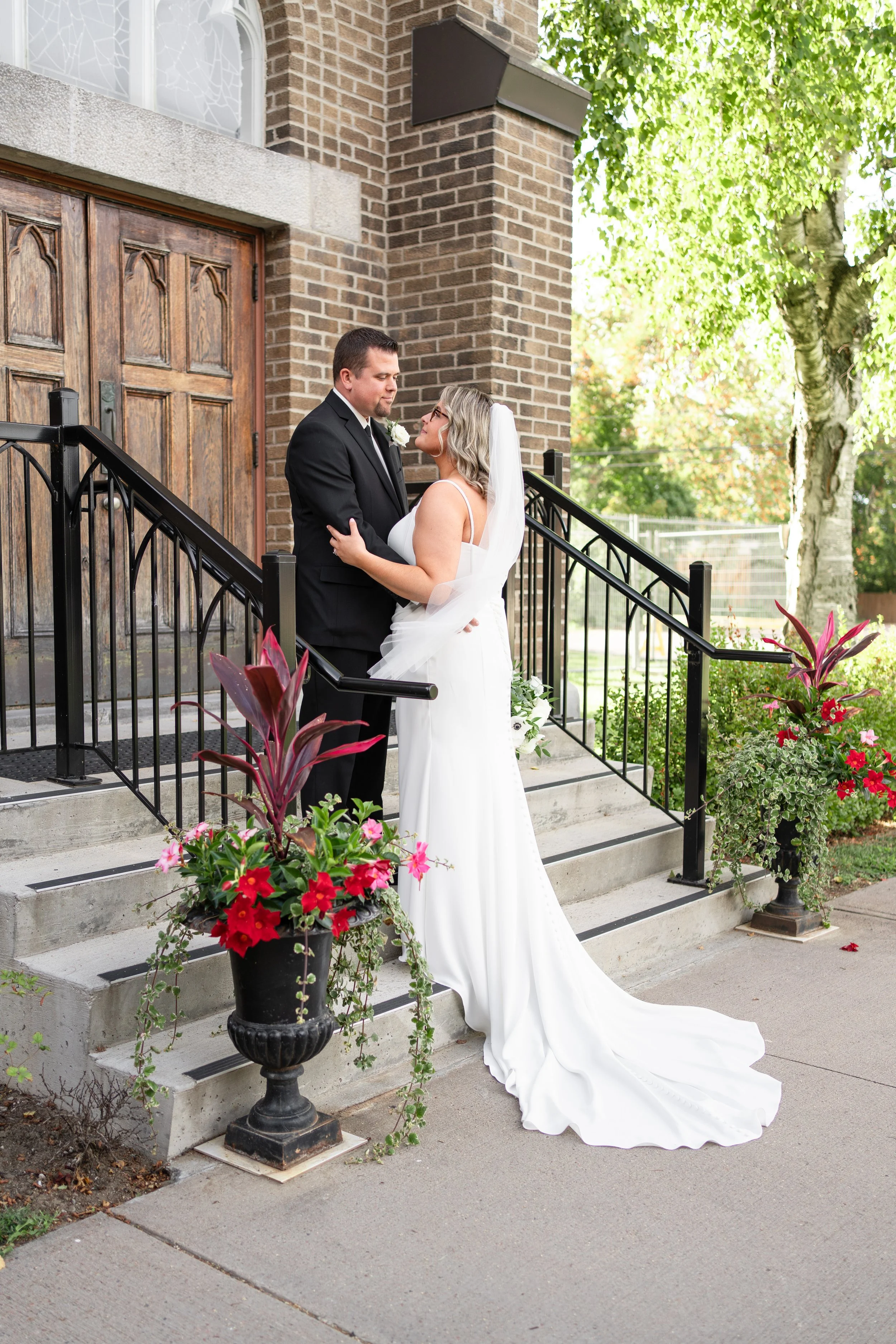 A bride and groom standing closely on the steps outside a church, gazing into each other's eyes, with flowers in black urns on either side of the staircase. church in Pembroke, Ontario