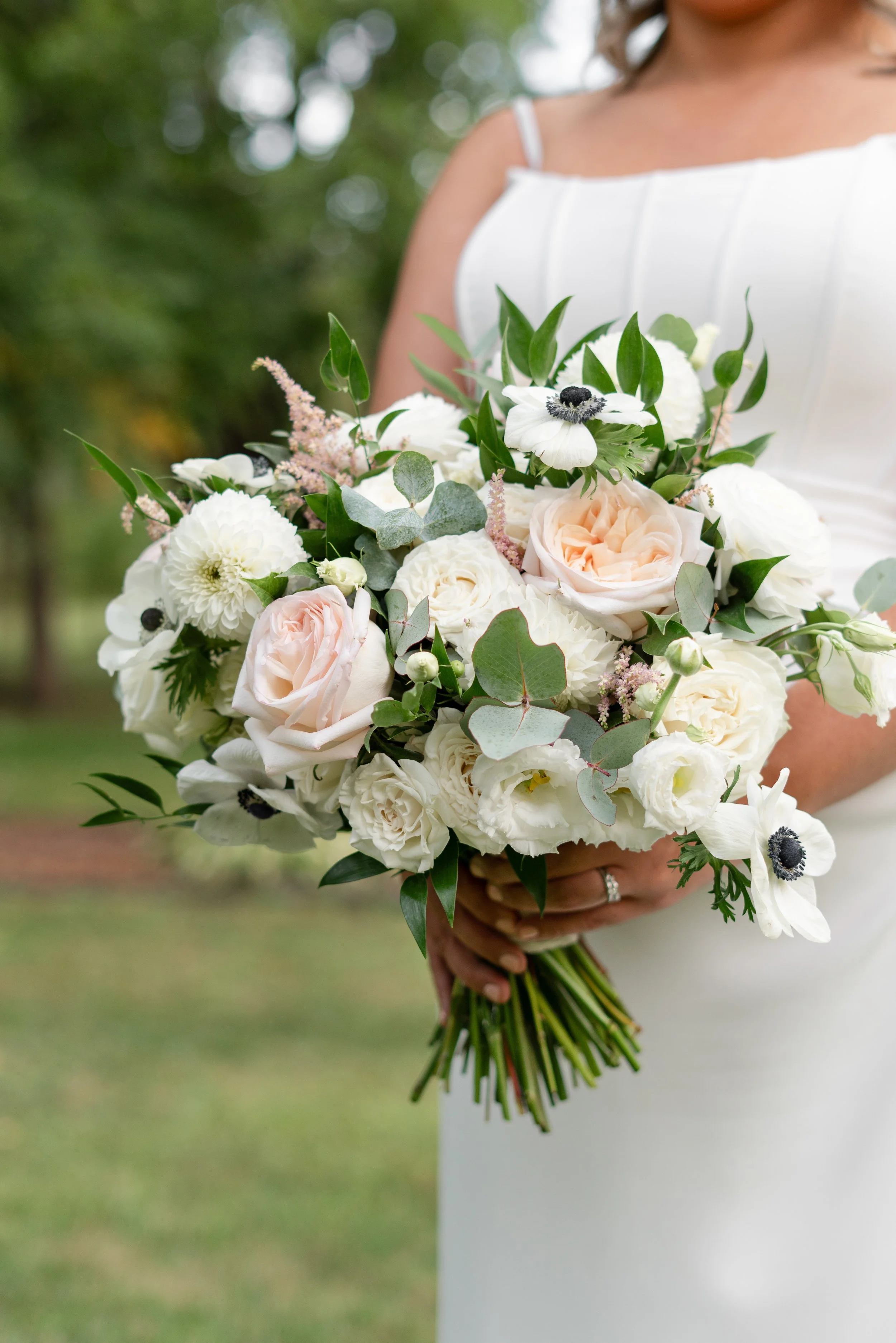 A bride holding a large bouquet of white and blush pink roses, white anemones with black centers, small white dahlias, and assorted greenery. 