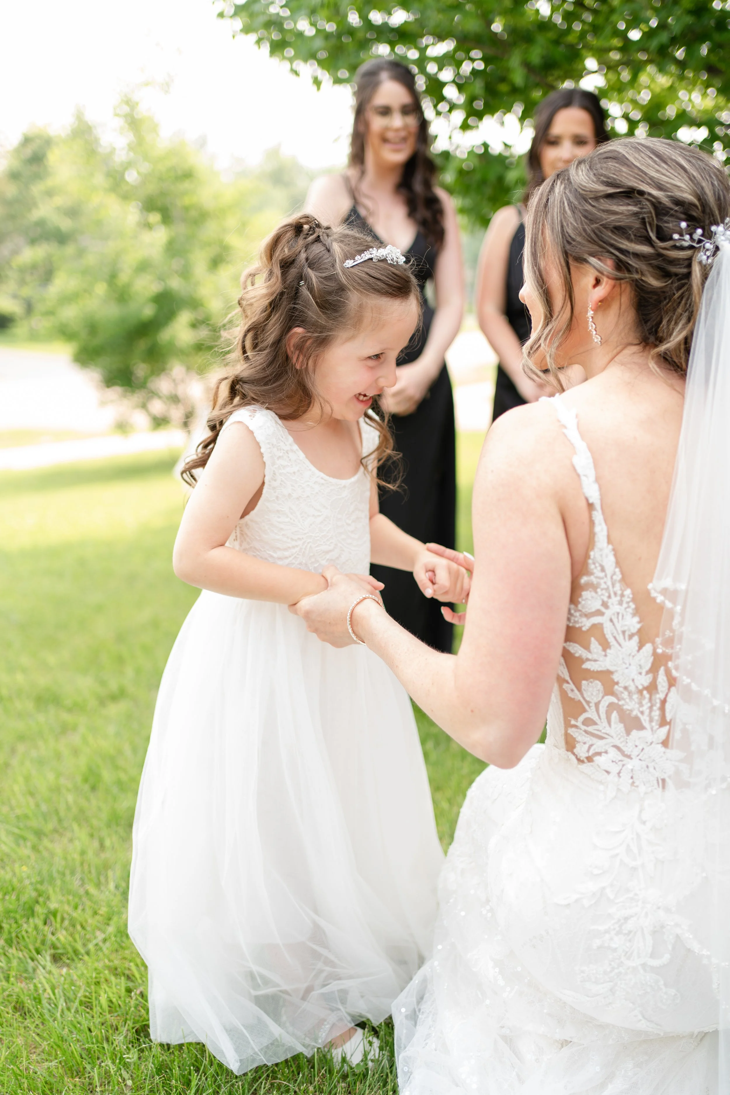 A young girl in a white dress holds hands with a bride in a wedding dress while smiling. In the background, bridesmaids, look on and smile. 