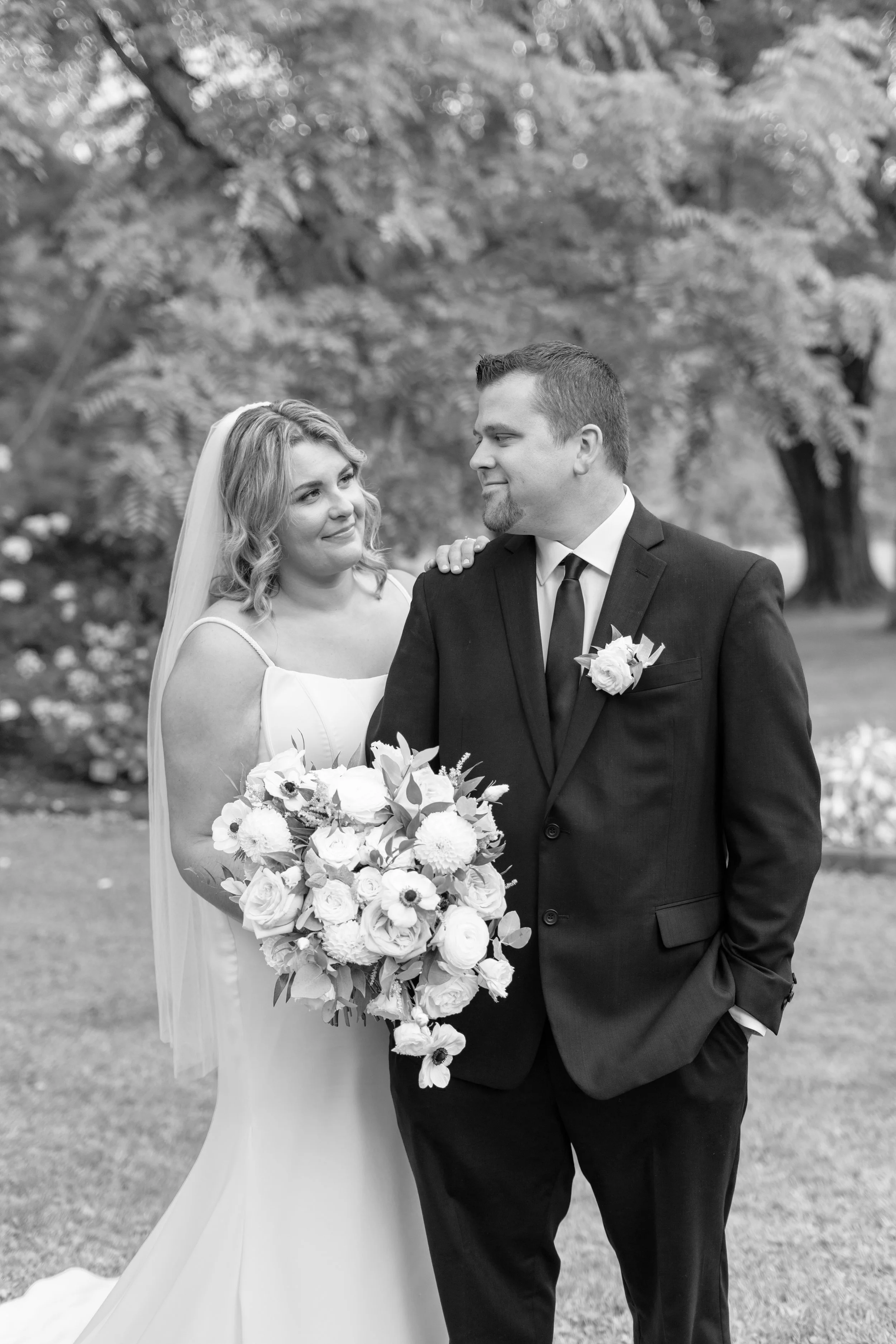 Black and white photo of a bride and groom standing outdoors, looking at each other. The bride is holding a large bouquet and has her hand on the groom's shoulder. 