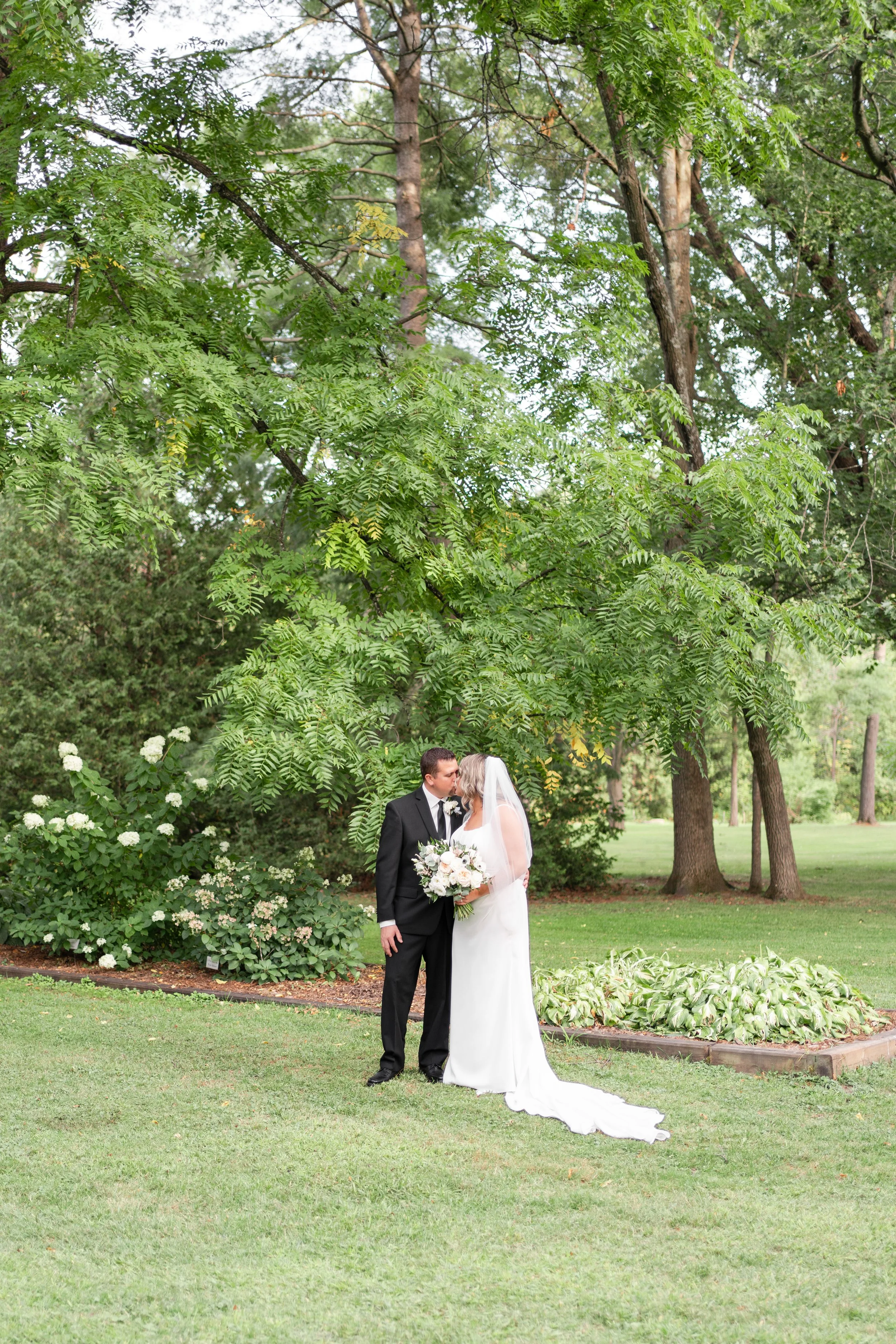 A bride and groom standing close together, kissing, outdoors in a lush garden with large trees and green foliage, during their wedding. Pemrboke park, Ontario.
