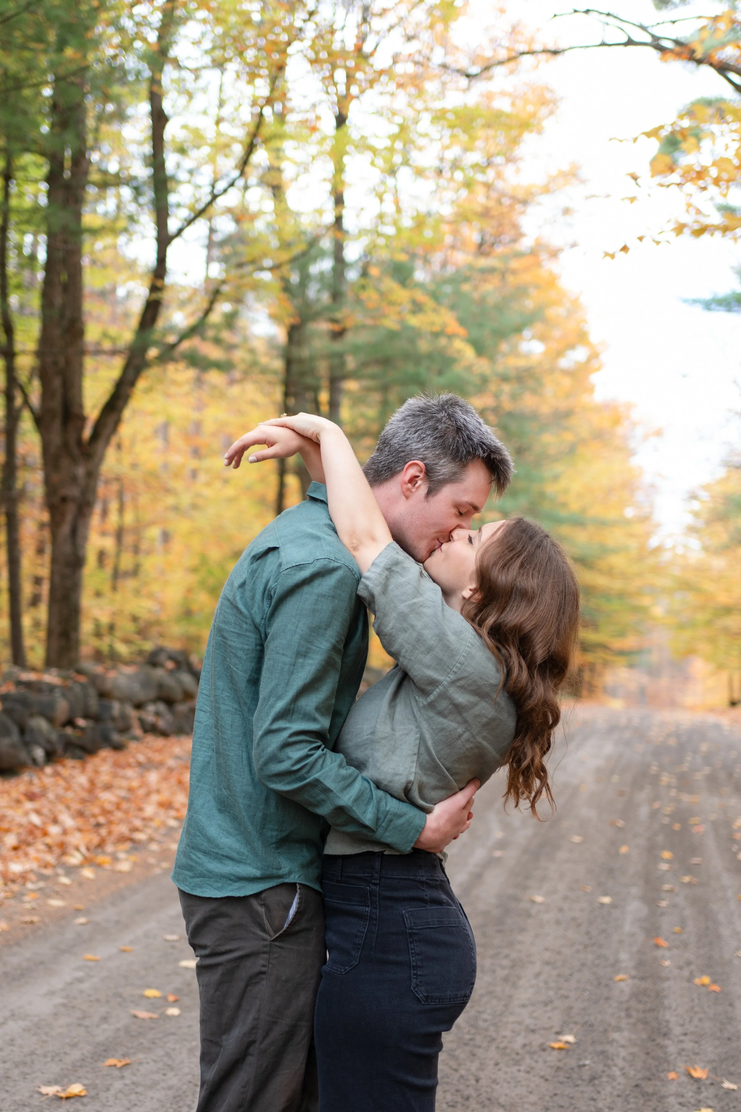 A couple kissing in a forest during fall, with leaves on the trees and the ground.