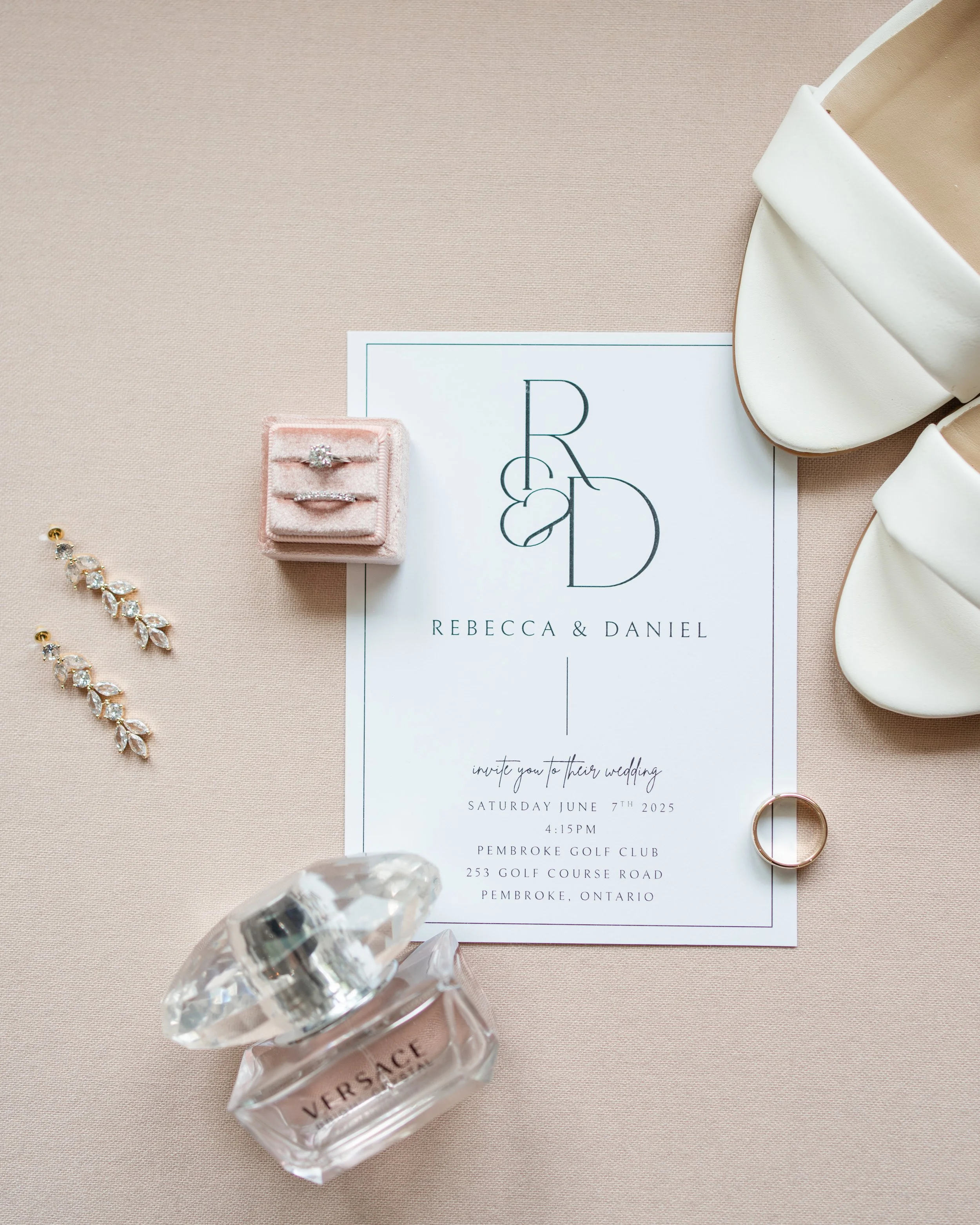 Wedding invitation surrounded by jewelry, perfume, shoes, and a ring on a pink surface. flat lay