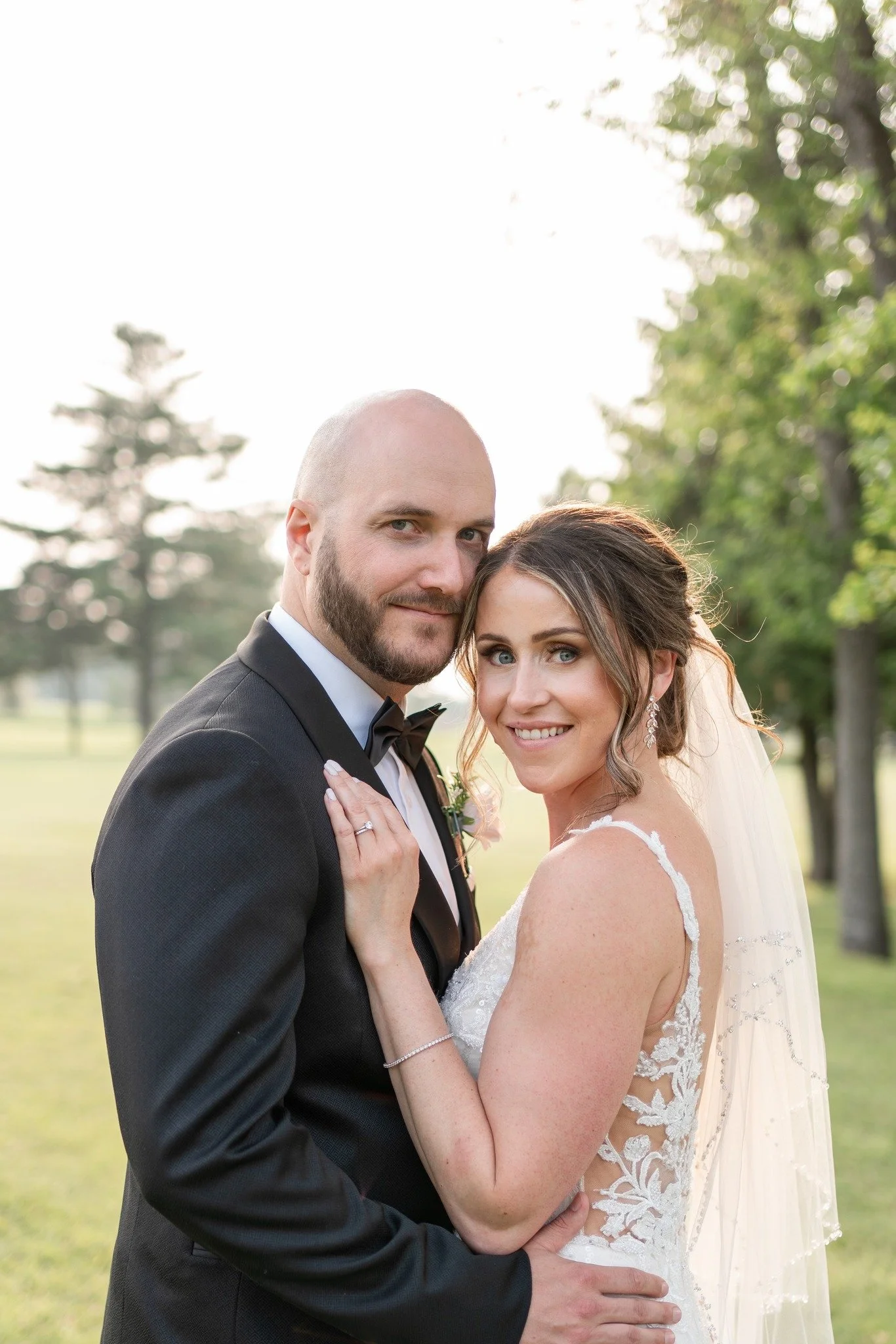 A newlywed couple standing closely outdoors, with a man in a black tuxedo and bow tie and a woman in a white lace wedding dress with a veil, smiling at the camera.