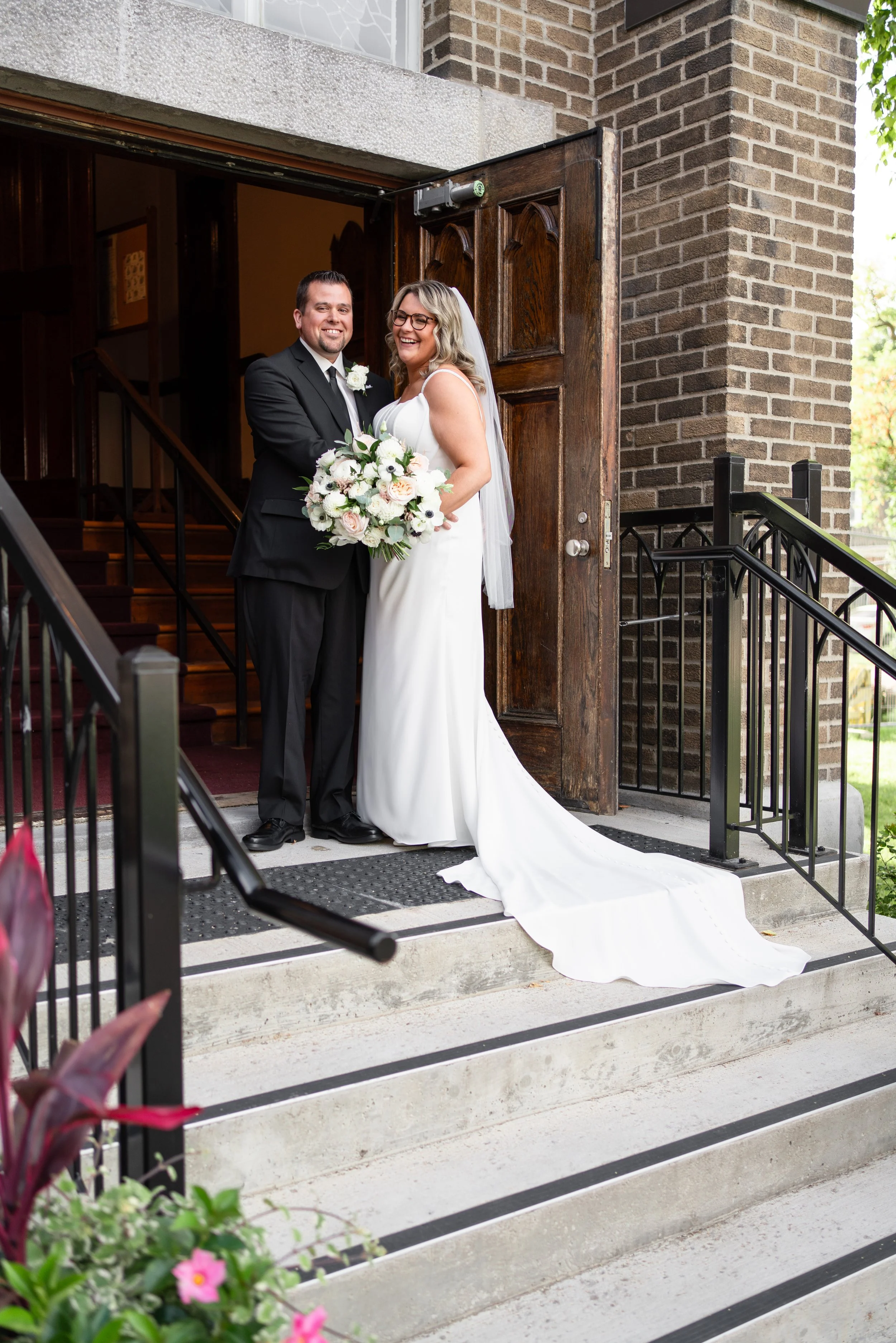Bride and groom standing at church entrance, smiling, with the bride holding a bouquet of white and pink flowers, and the groom in a black suit, in front of wooden church doors. Pemrboke church, Ontario