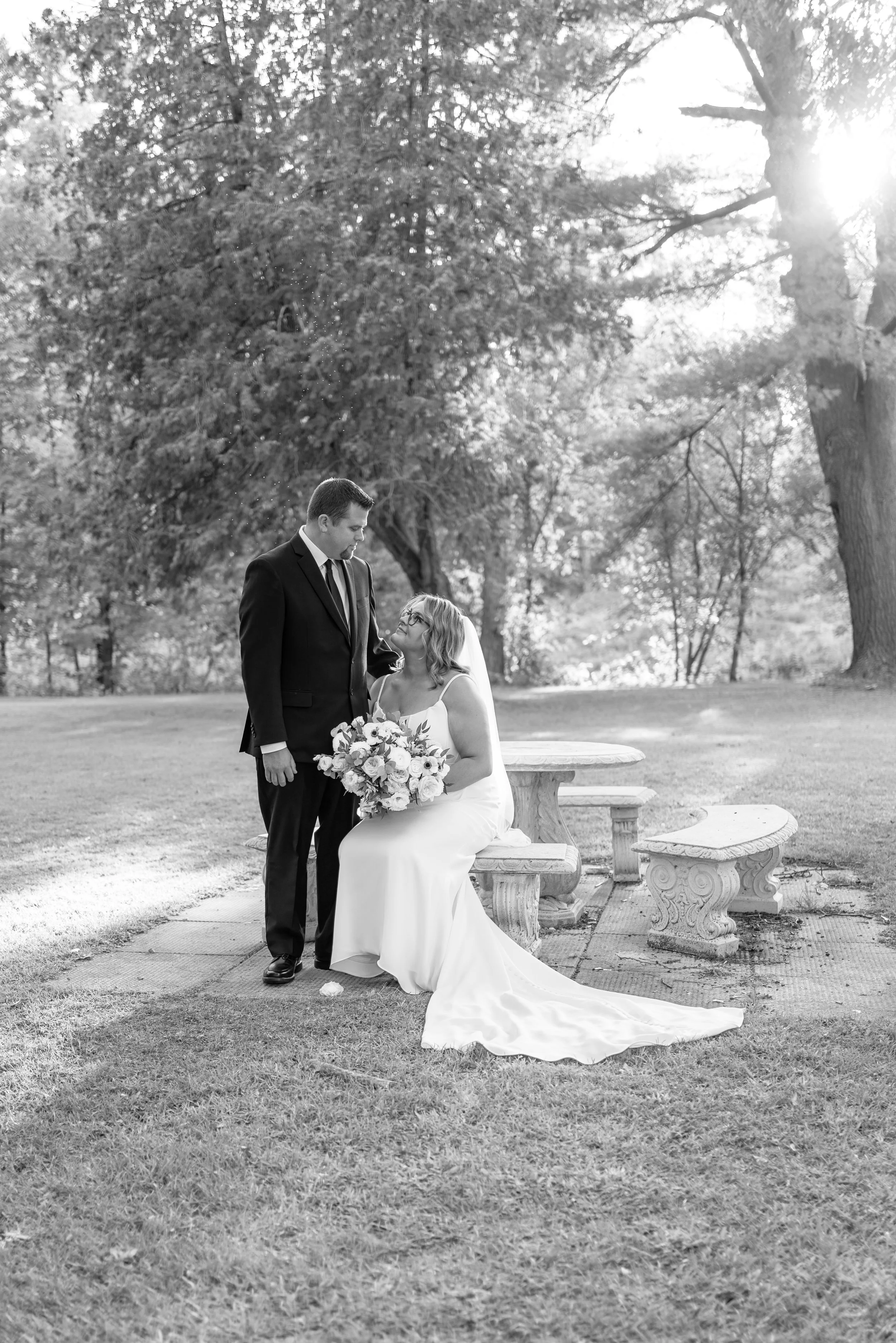 A bride sitting on a bench holding a bouquet of flowers, and a groom standing beside her, looking at her in a park with trees and sunlight in the background, in black and white. Pembroke wedding in Ontario