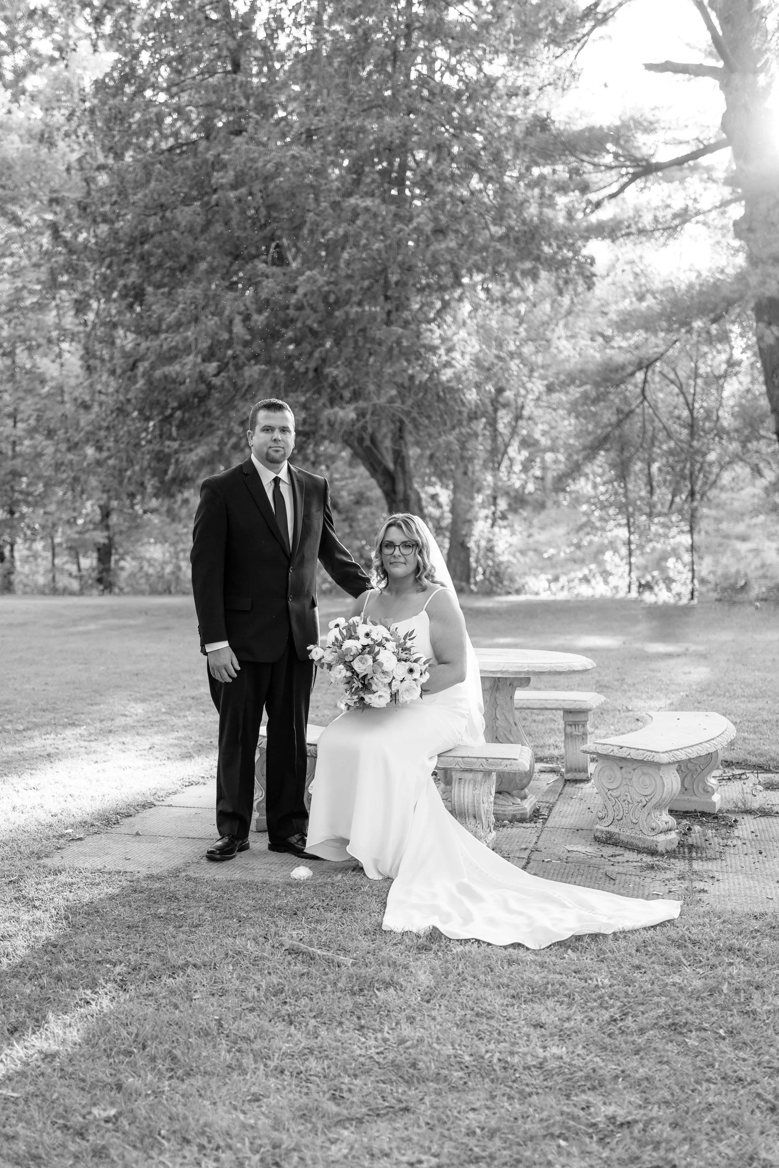 A bride in a white wedding dress holding a bouquet of flowers, sitting on a stone bench, with a groom in a black suit and tie standing beside her in a park with trees and sunlight. Pemrboke park