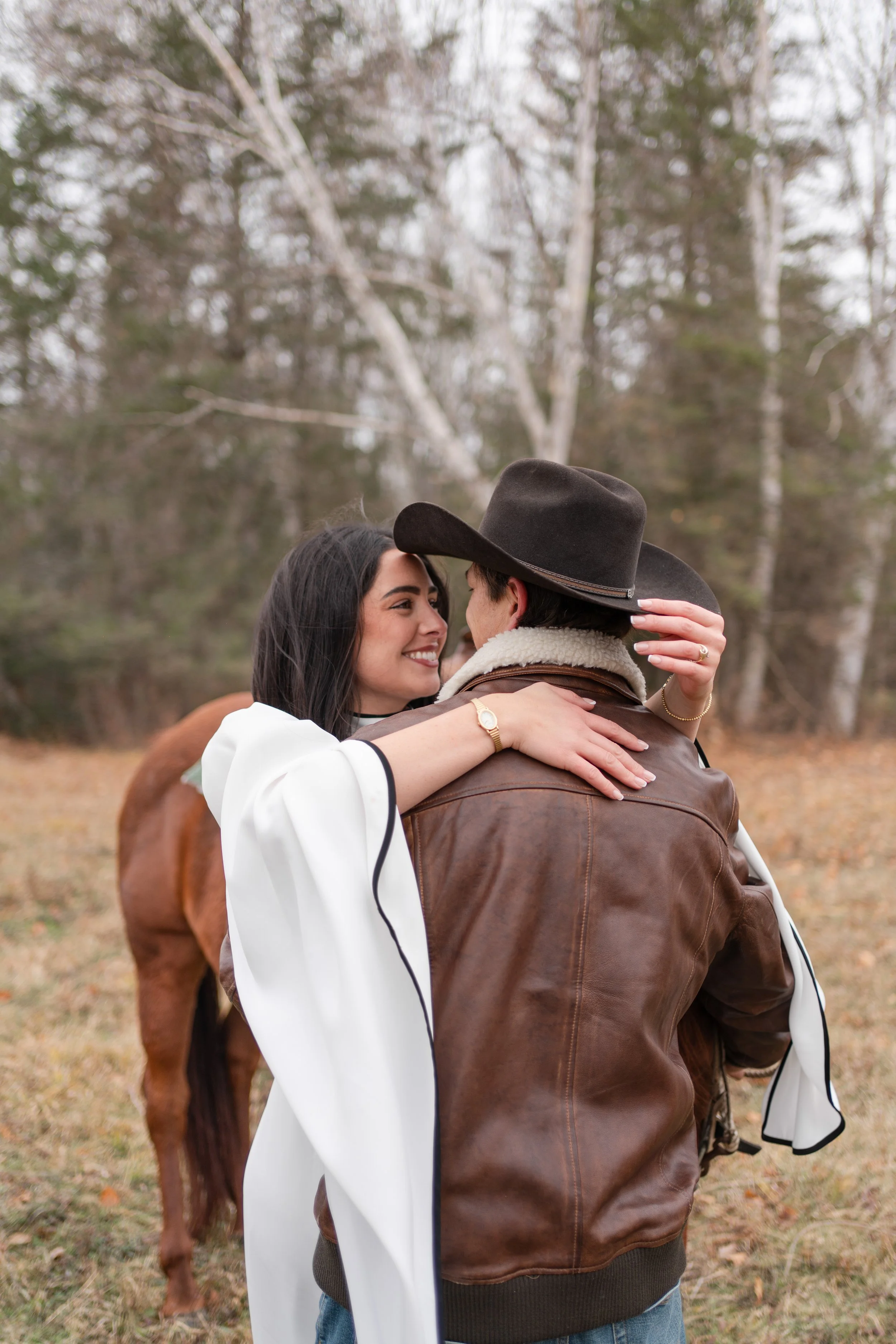 A woman and a man embrace outdoors in a fall setting, with bare trees and a horse in the background.