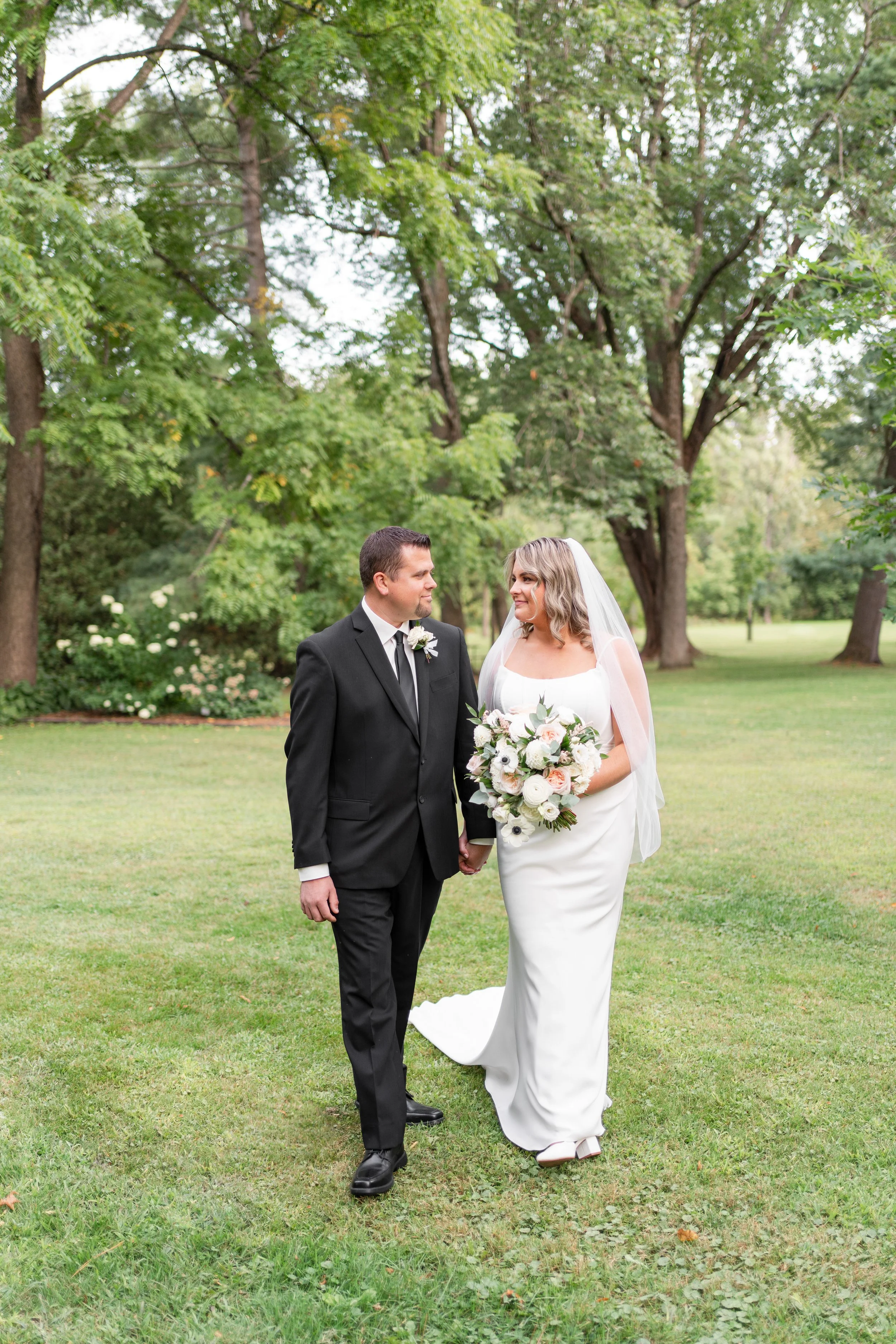 Bride and groom holding hands in a park during wedding, groom in black suit and tie, bride in white dress with veil, holding bouquet, trees in background. Pemrboke, Ontario