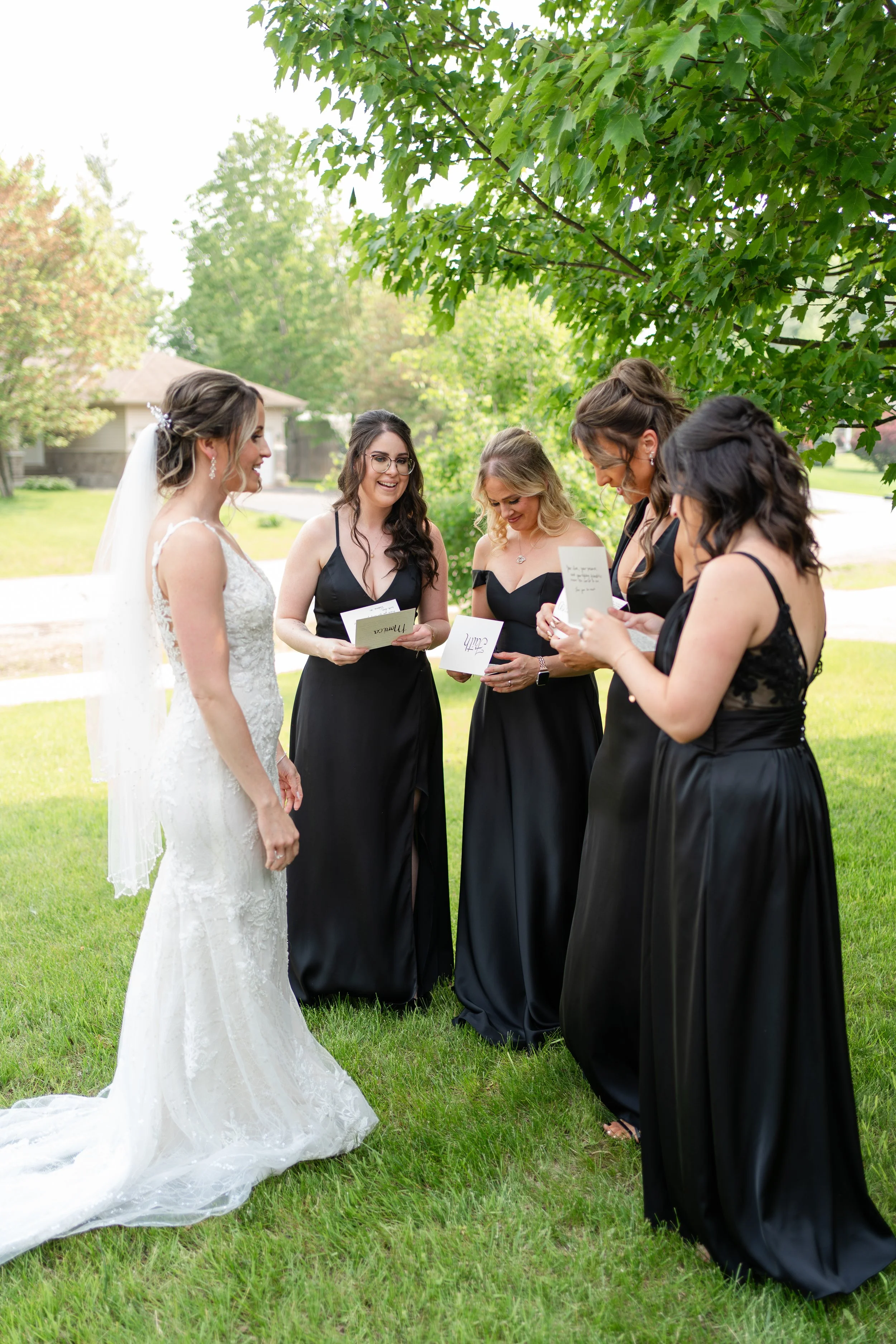 Bride in a white lace wedding gown with a veil standing outdoors with five bridesmaids in black dresses, holding greeting cards, under a leafy tree with a grassy lawn in the background.