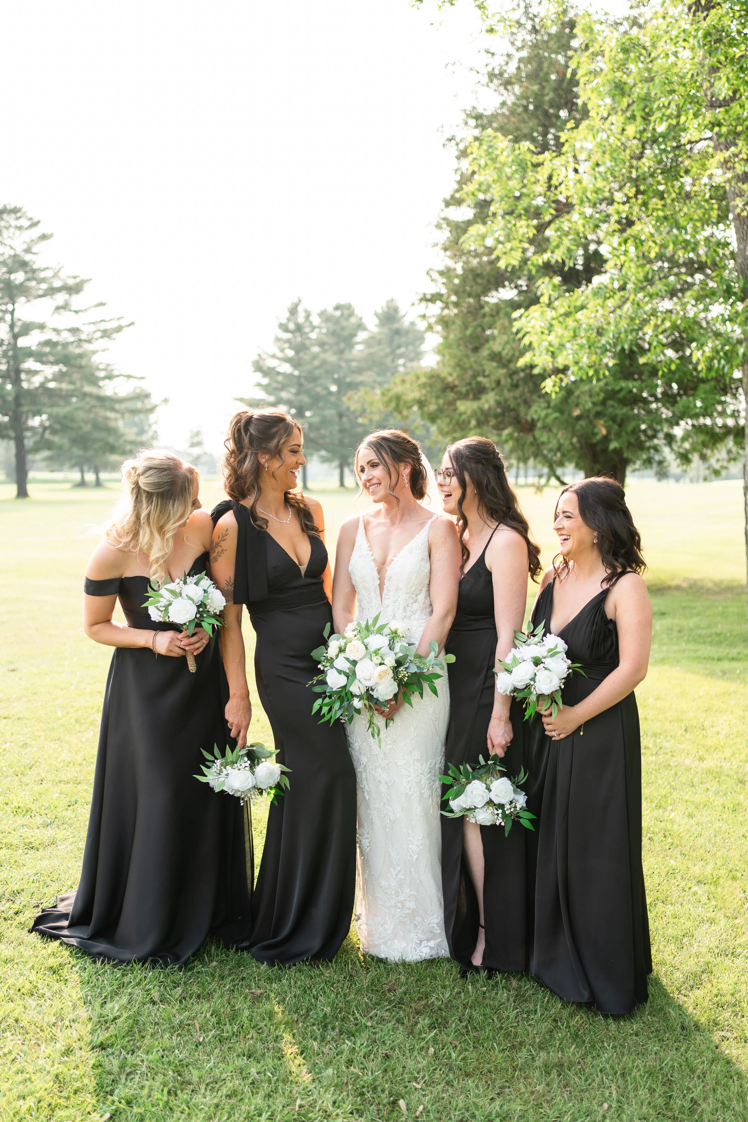 A bride in a white lace wedding dress with four bridesmaids in black dresses holding white bouquets, standing on a grassy field with trees in the background. Wedding in Pembroke, Ontario Pembroke Golf Club