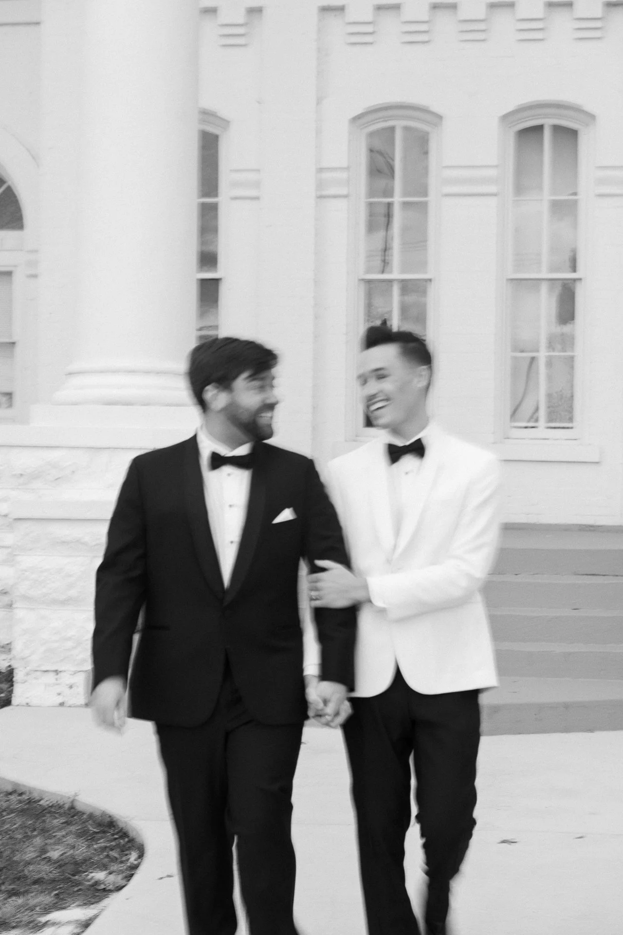 Two men in tuxedos, holding hands, smiling while walking outside a building with large columns and windows.