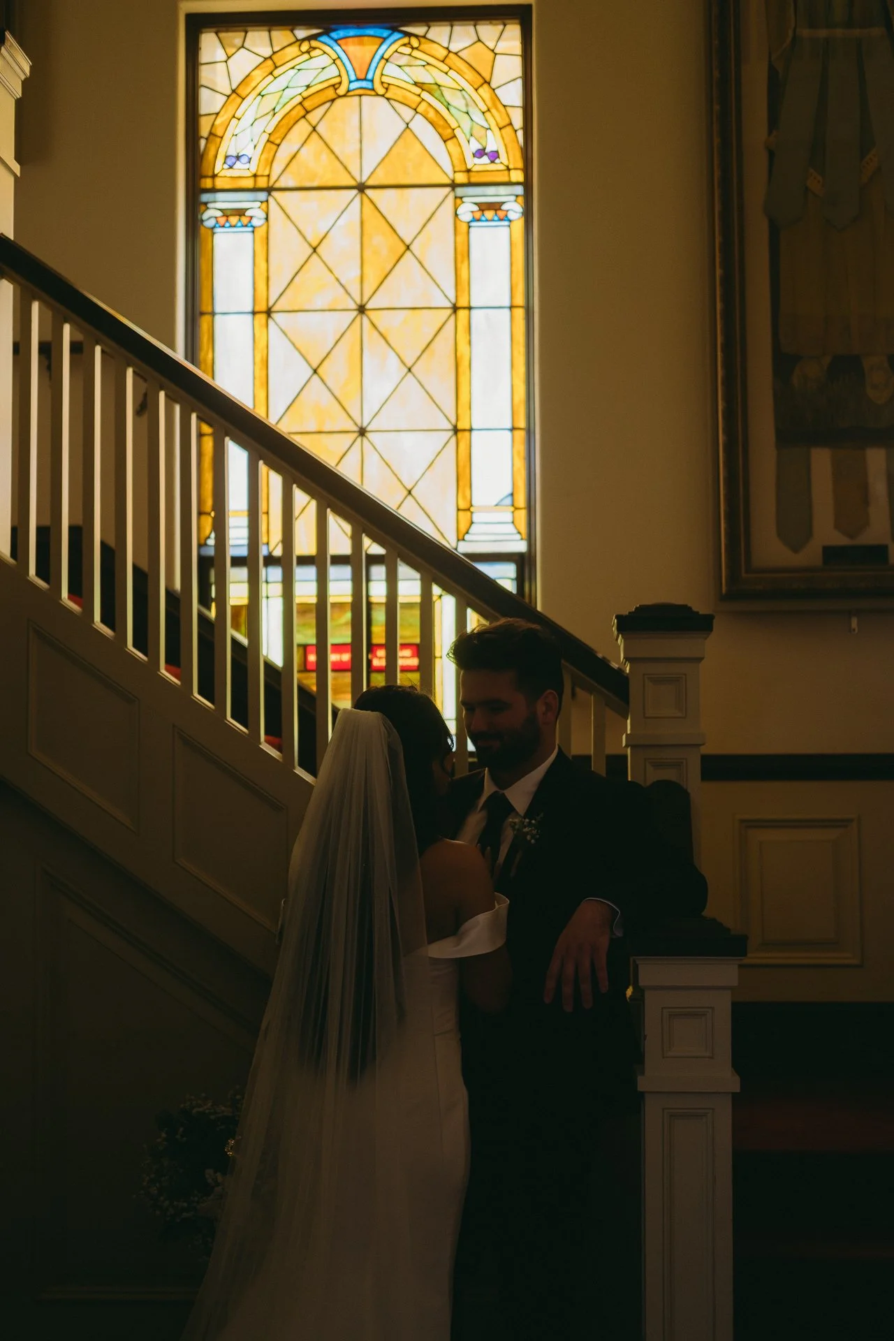 A bride and groom in wedding attire are silhouetted inside a church near a staircase, with a large stained glass window in the background.
