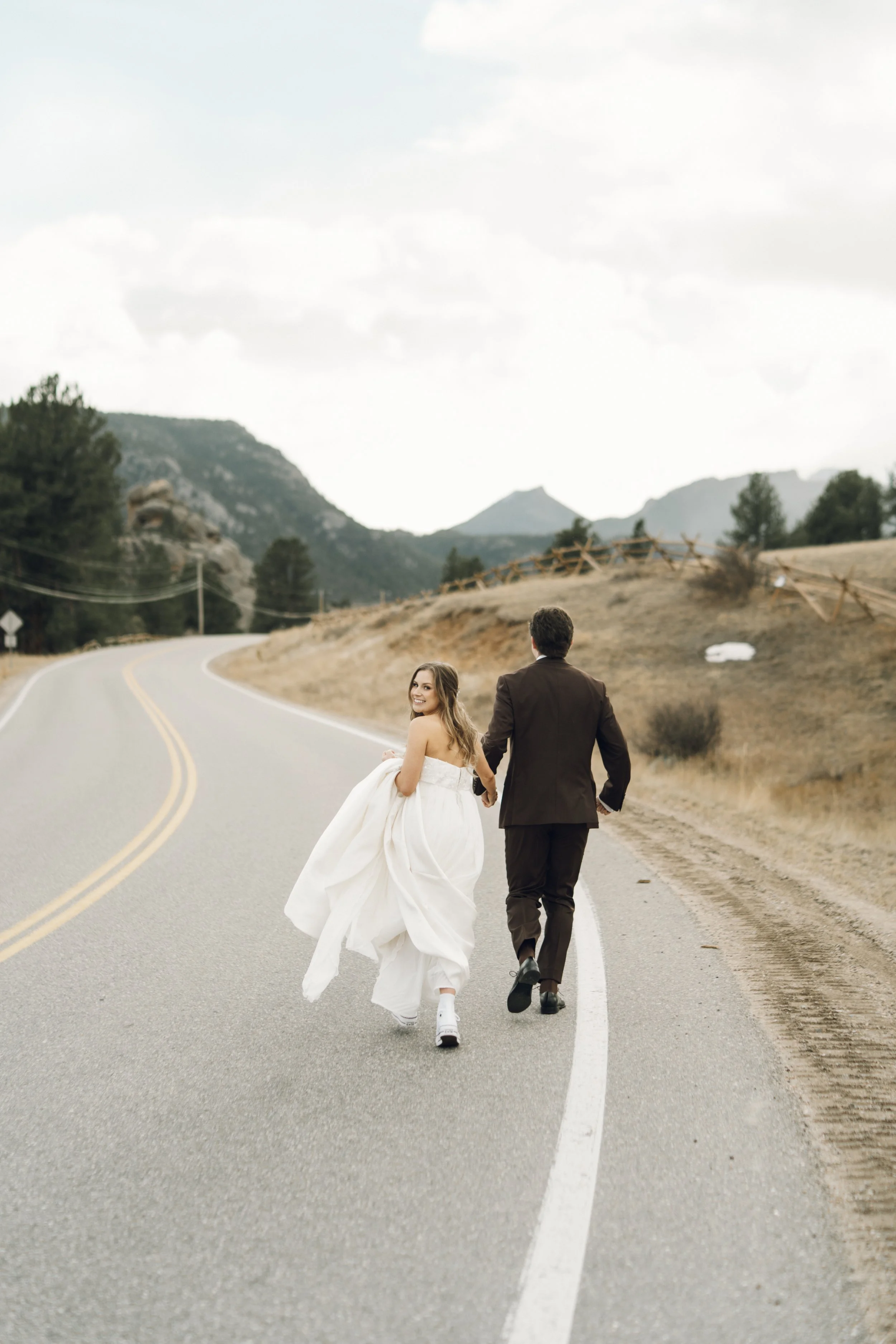 A couple in wedding attire walking hand in hand on a winding road in a rural mountainous area, with the bride smiling at the camera.