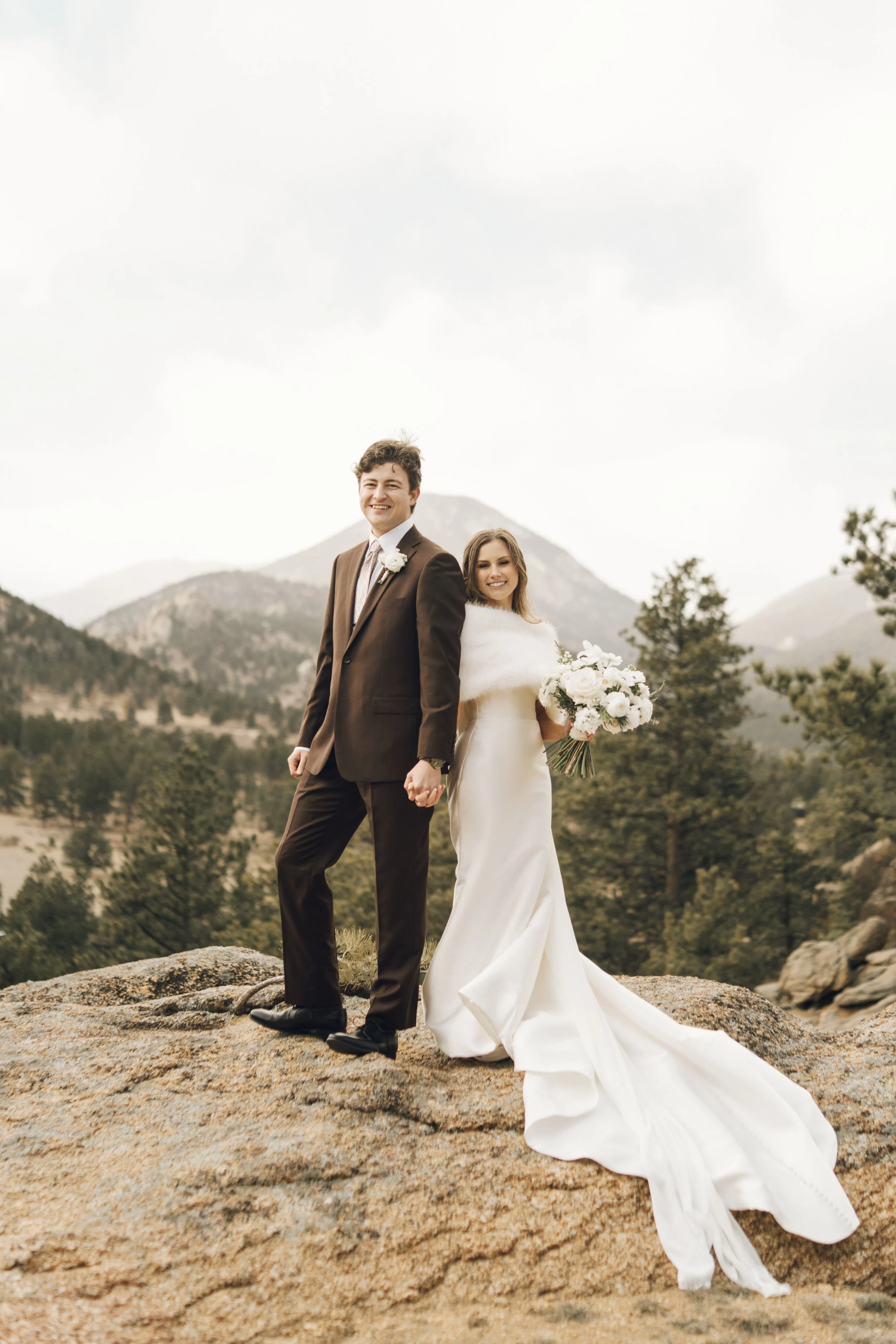 A newlywed couple standing outdoors on a rocky terrain with mountain and forest background, smiling and holding hands. The bride is in a white wedding dress and holding a bouquet, the groom is in a dark suit.