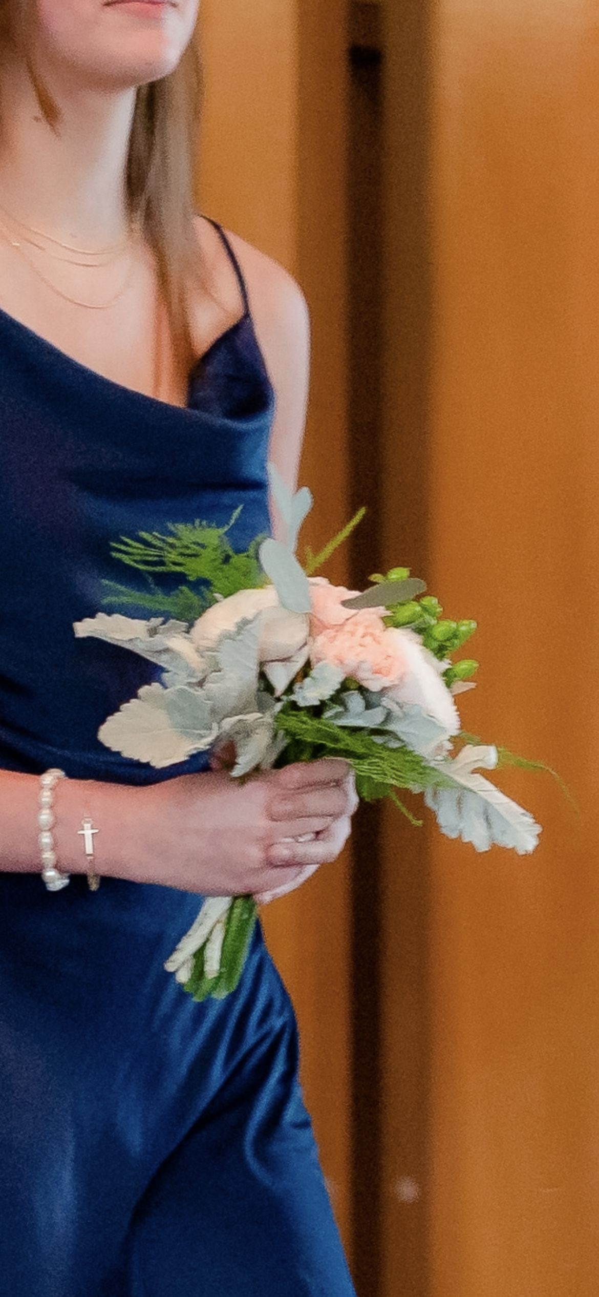 A woman dressed in navy blue satin gown holding a bouquet of pink and white flowers with green foliage, visible jewelry including a pearl bracelet with a cross charm and multiple delicate gold necklaces.