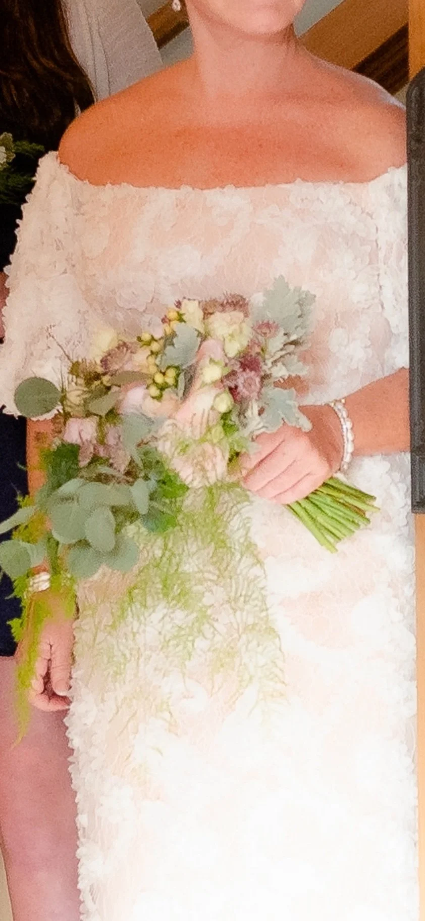 A woman in a white off-the-shoulder dress holding a bouquet of flowers, wearing a bracelet on her right wrist.