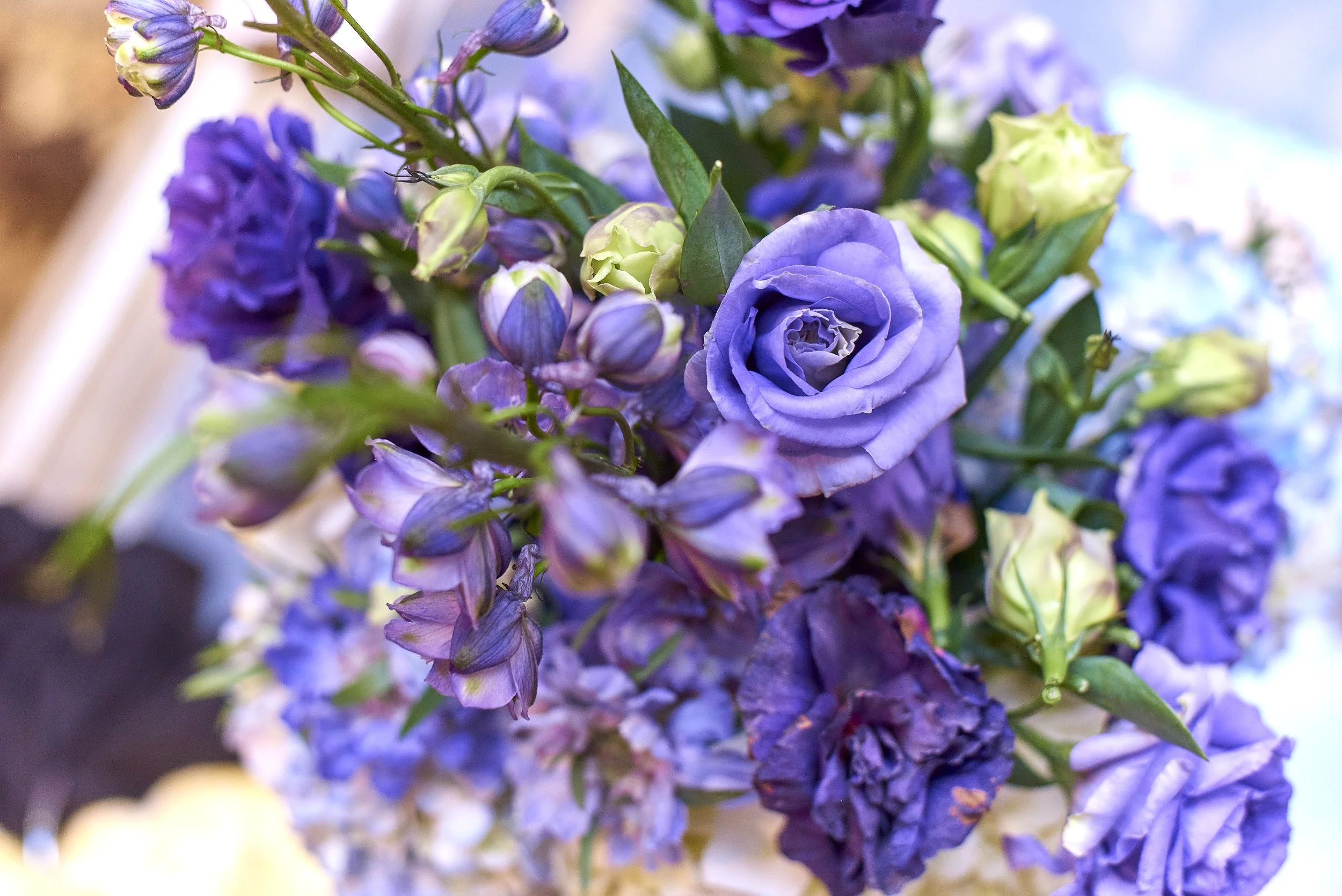 Close-up of a bouquet with purple, lavender, and pale yellow flowers, including roses and others, with green leaves.