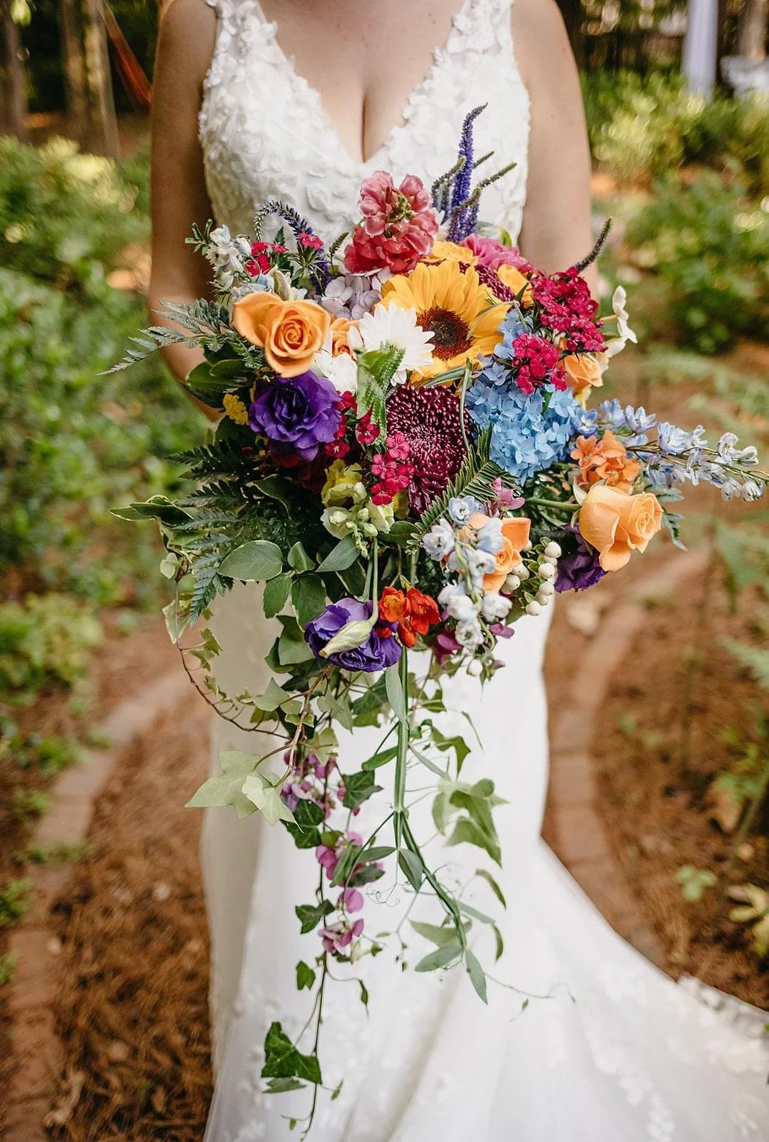 Woman in white wedding dress holding a cascading bouquet of colorful flowers, including sunflowers, roses, hydrangeas, and delphiniums, outdoors.