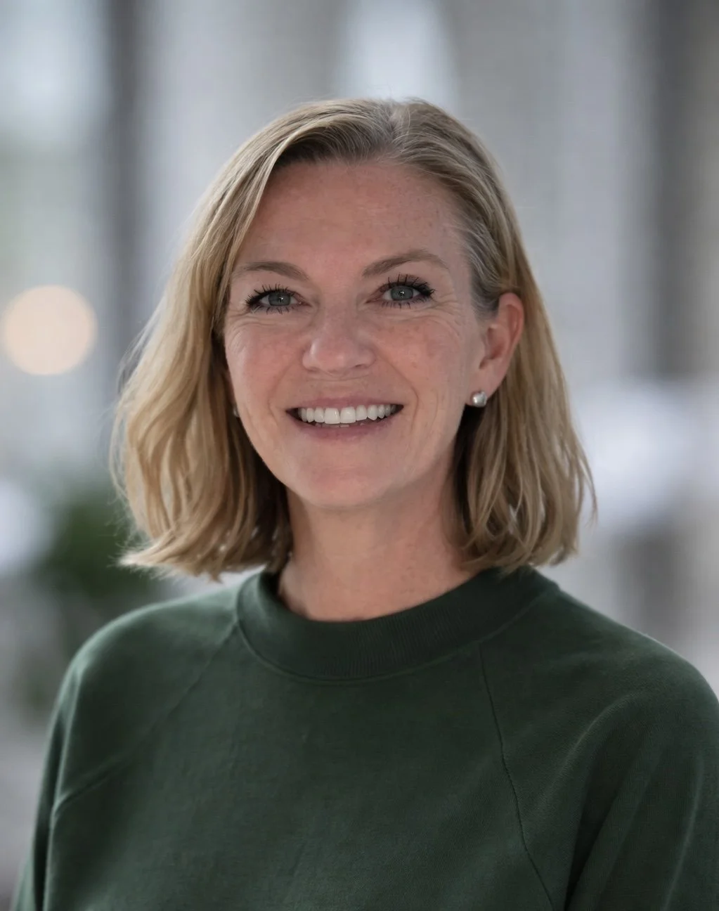 A smiling woman with blonde hair wearing a dark green top in an indoor setting.