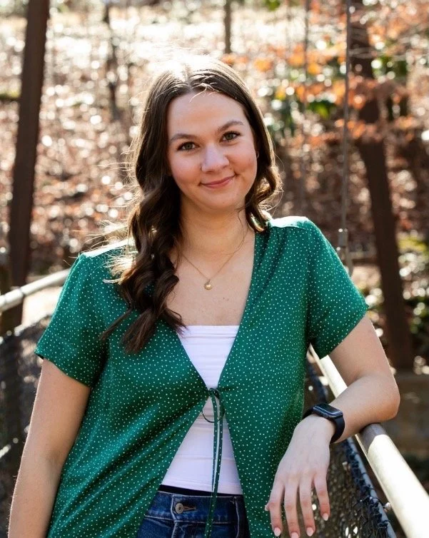 A young woman with long brown hair, wearing a green polka-dot blouse over a white top, standing outdoors on a bridge during autumn.