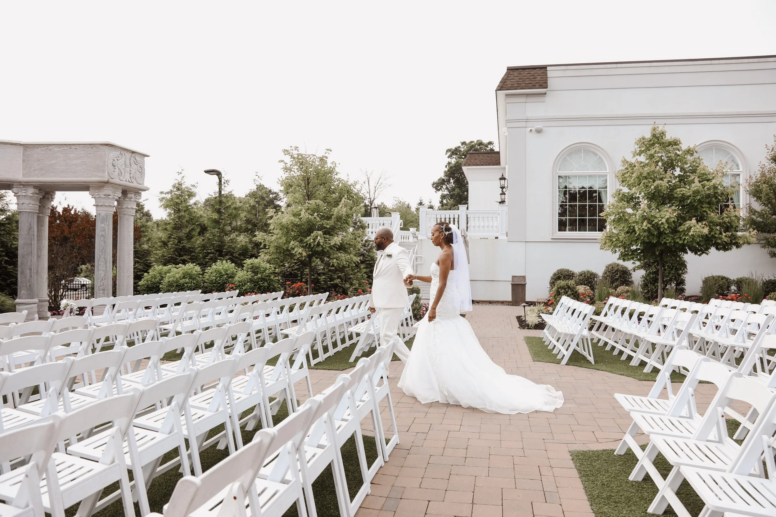 A bride and groom walking hand in hand on an outdoor wedding aisle surrounded by white chairs, with trees and a white building in the background.