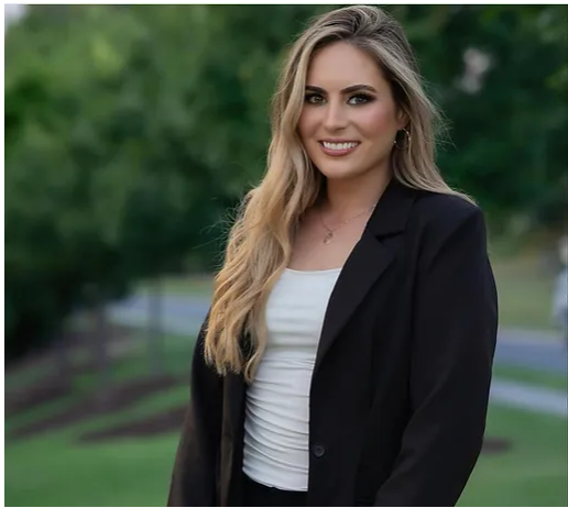 Portrait of a young woman with long blonde hair, wearing a black blazer over a white top, smiling outdoors with blurred green trees in the background.