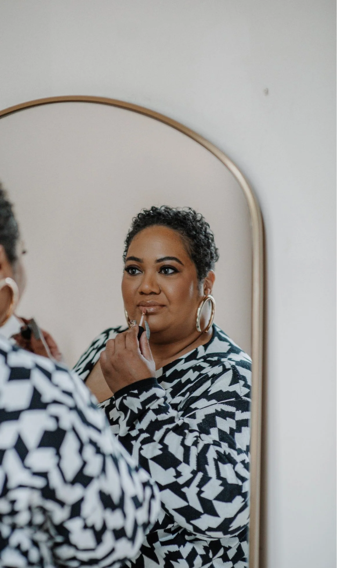A woman with short, curly hair looking into a mirror, applying lip gloss.