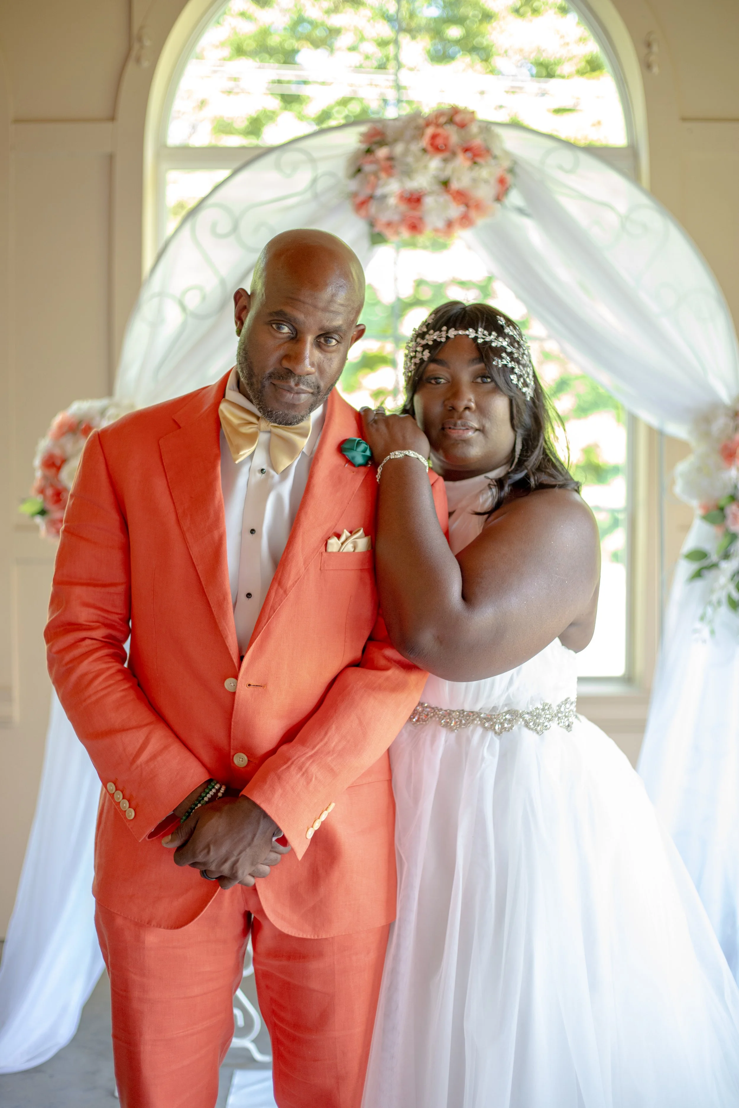 A couple dressed in formal wedding attire standing underneath a decorated archway with flowers and white drapery, in front of a window with trees outside.
