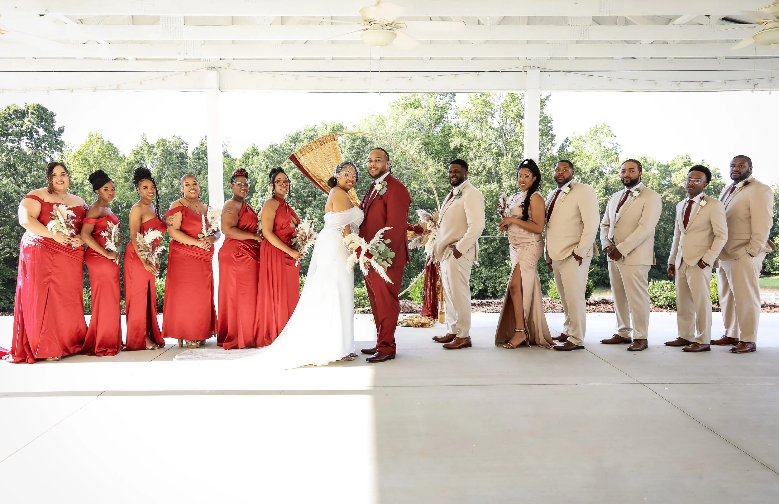 Wedding party with bride and groom in center, bridesmaids in red dresses on left, groomsmen in beige suits on right, standing under open pavilion with green trees in background.