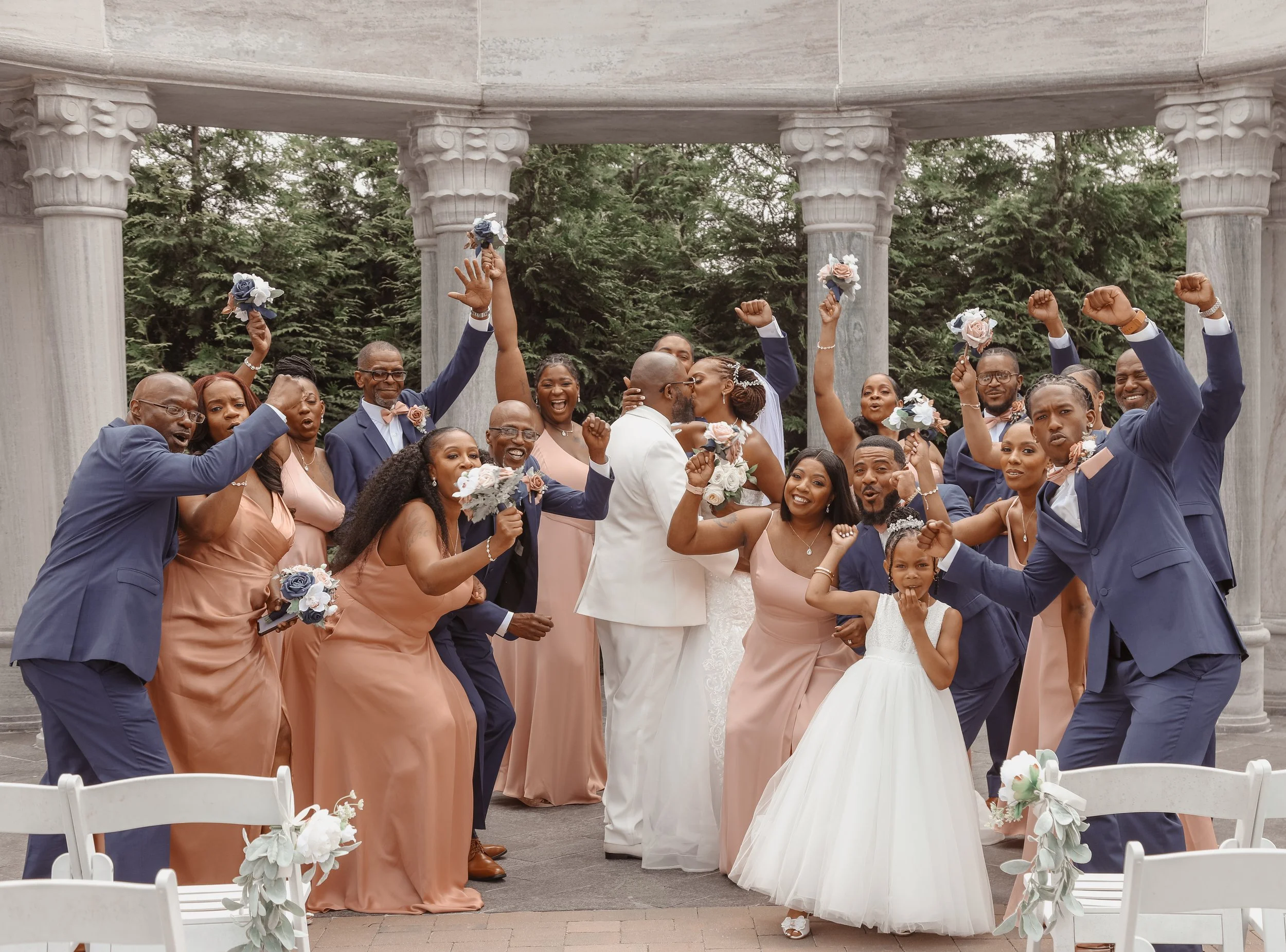 Celebrating a wedding outdoors with a large group of people, a bride and groom, and family and friends in formal attire, some holding bouquets, cheering and posing happily.