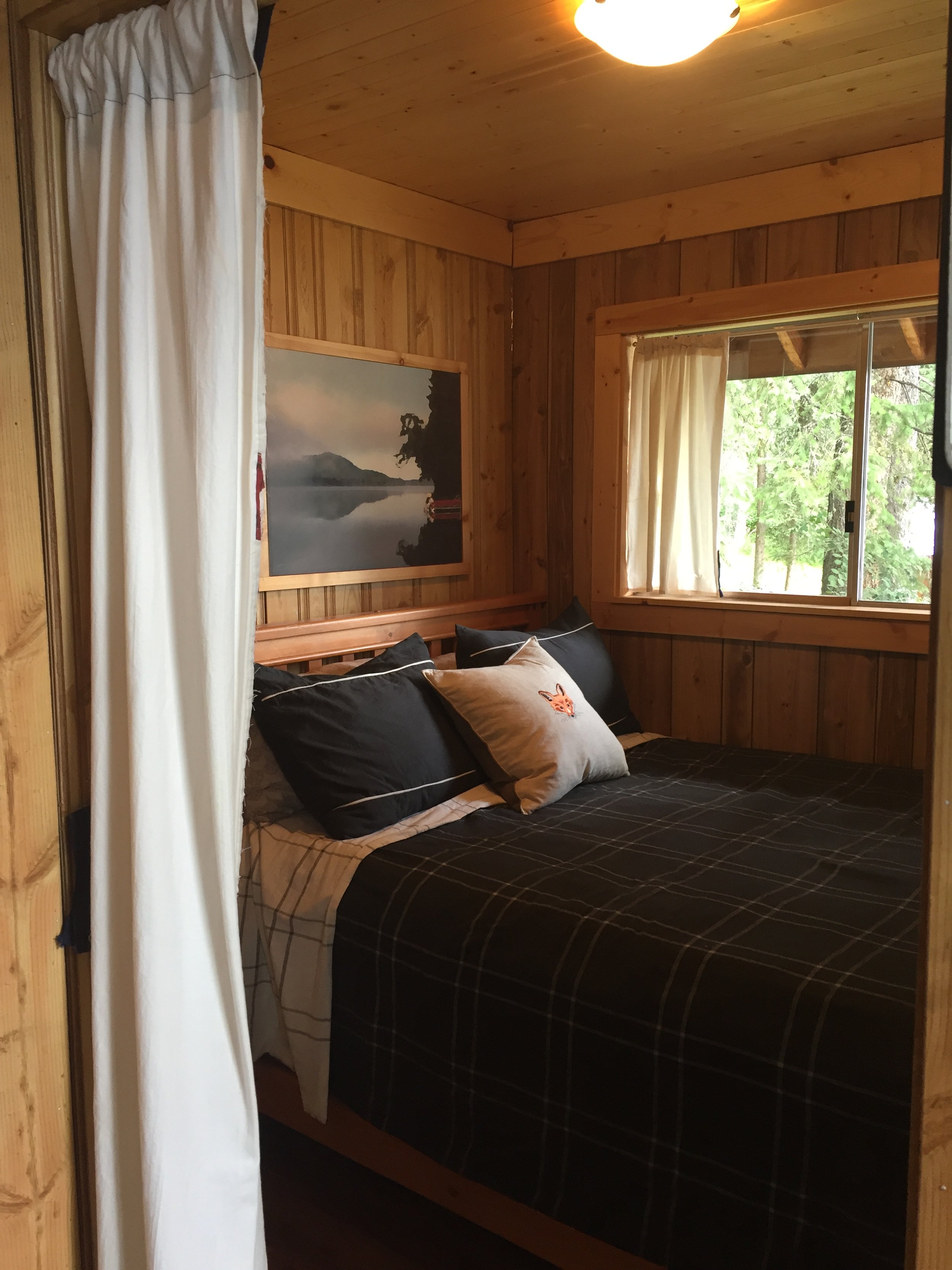 A peak into a tiny cabin bedroom that only fits a queen sized bed. There is a lakeview painting above the headboard. The window is extra large and it gets reframed in the renovation to a smaller window.