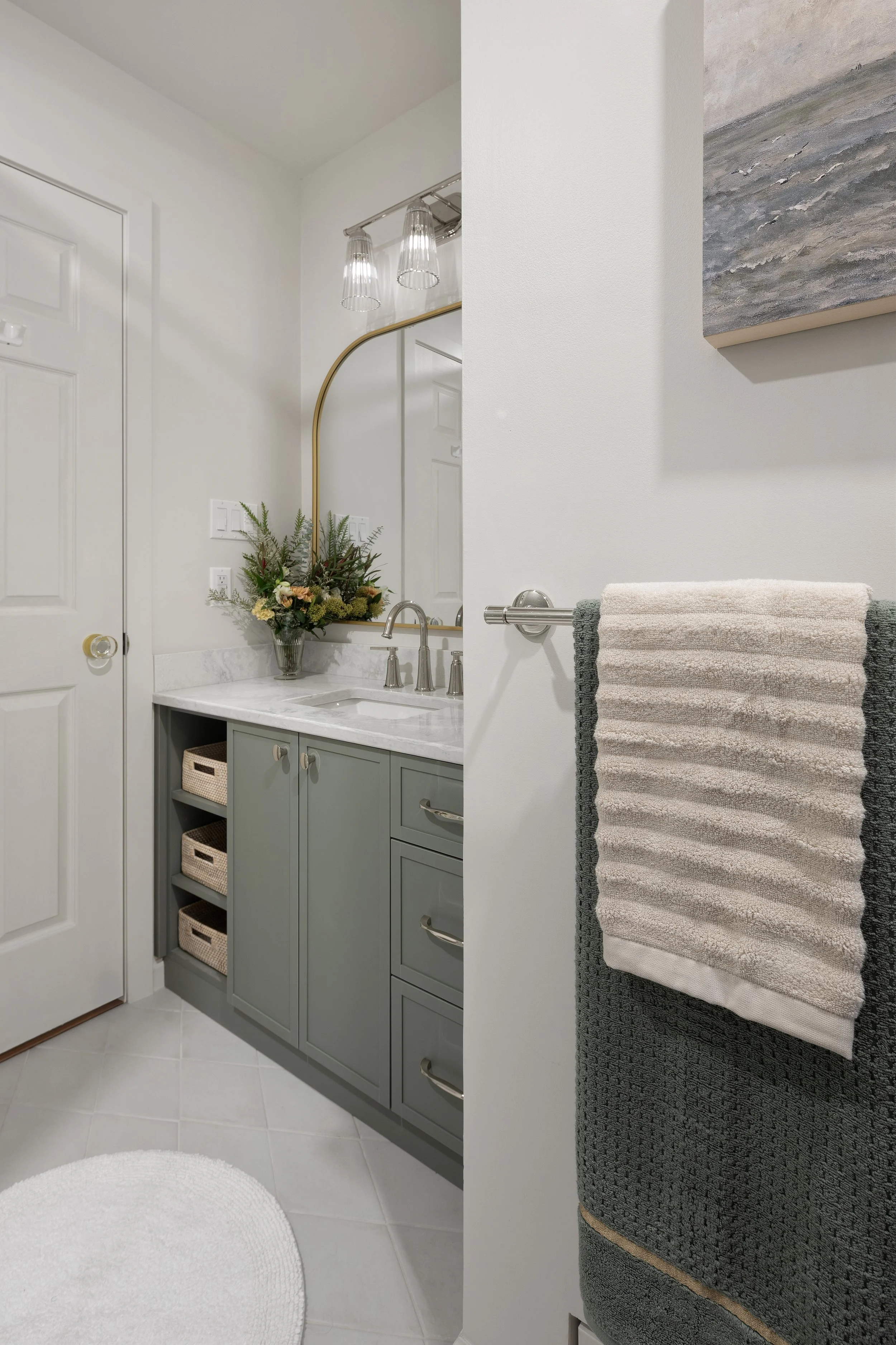 Renovated bathroom is crisp with soft green vanity. The floor is white Ossidi diamond tile from Olympia tile. A round white rug helps soften the space. A towel rack and art are in the foreground; the new vanity and mirror are in the background.