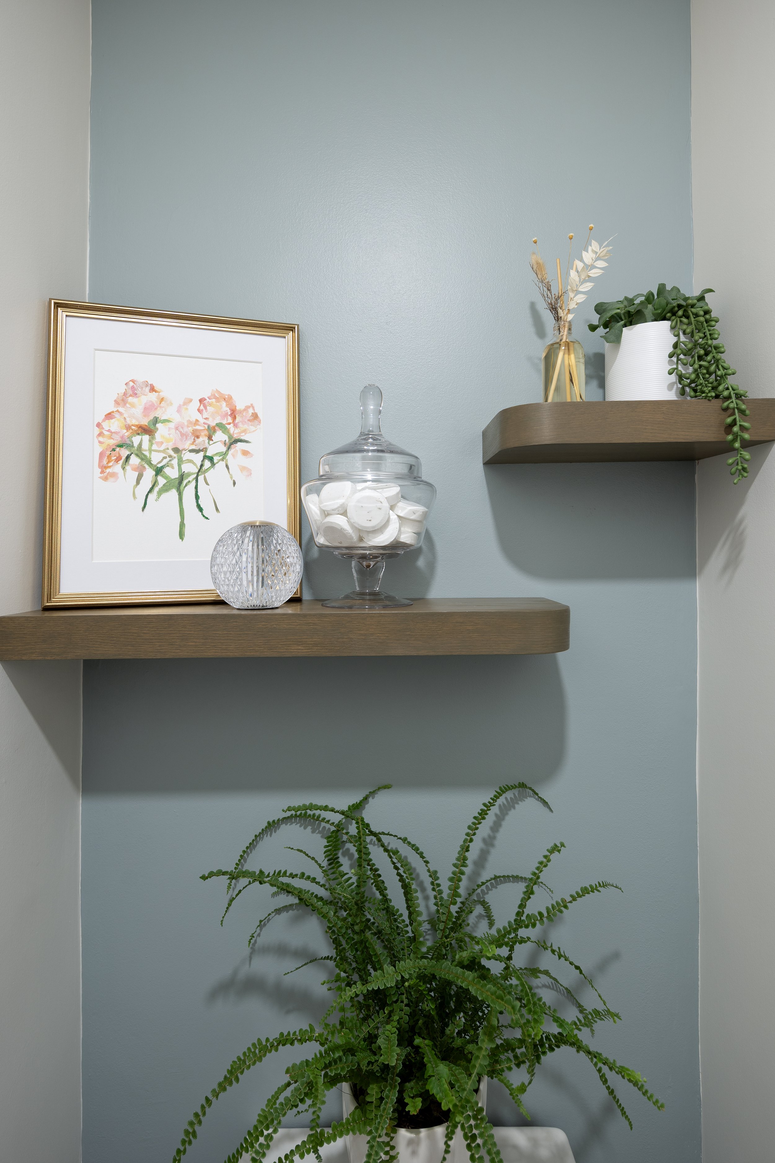 A narrow wall behind a toilet is freshened up with a painted blue wall in Sherwin Williams Eventide SW 9643 from the Emerald Designer Edition. Also custom shelves, plants and artwork make this hidden space more interesting and inviting.