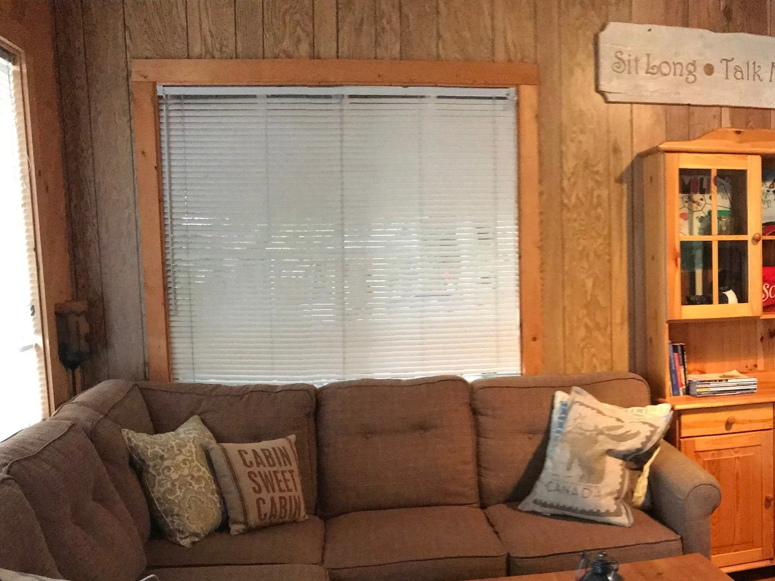 An old brown sectional sits in front of a 48" window with white mini blinds that are closed. The window seems large and takes up a lot of wall space but is not looking functional because the blinds are down.